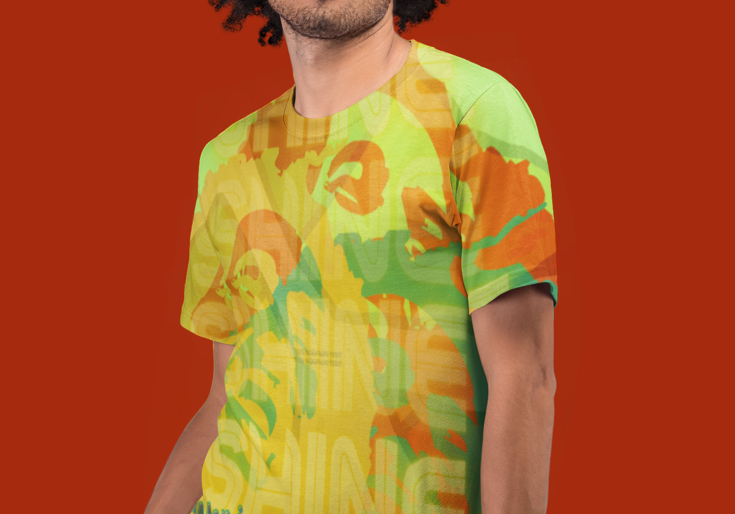 A man with curly hair stands against a solid red background, wearing a yellow and green patterned t-shirt with a quote that reads, "Man is a universe within himself. - Bob Marley." His hands are in his pockets.