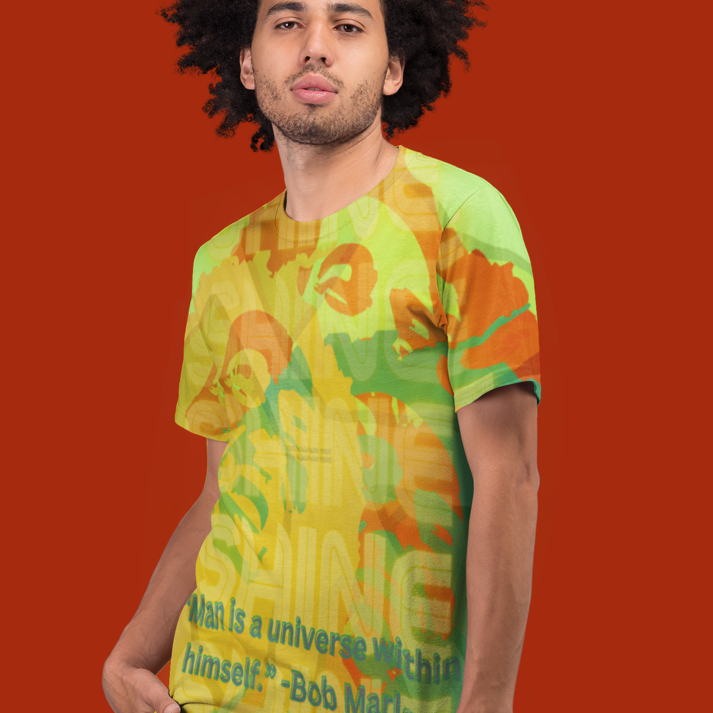 A man with curly hair stands against a solid red background, wearing a yellow and green patterned t-shirt with a quote that reads, "Man is a universe within himself. - Bob Marley." His hands are in his pockets.