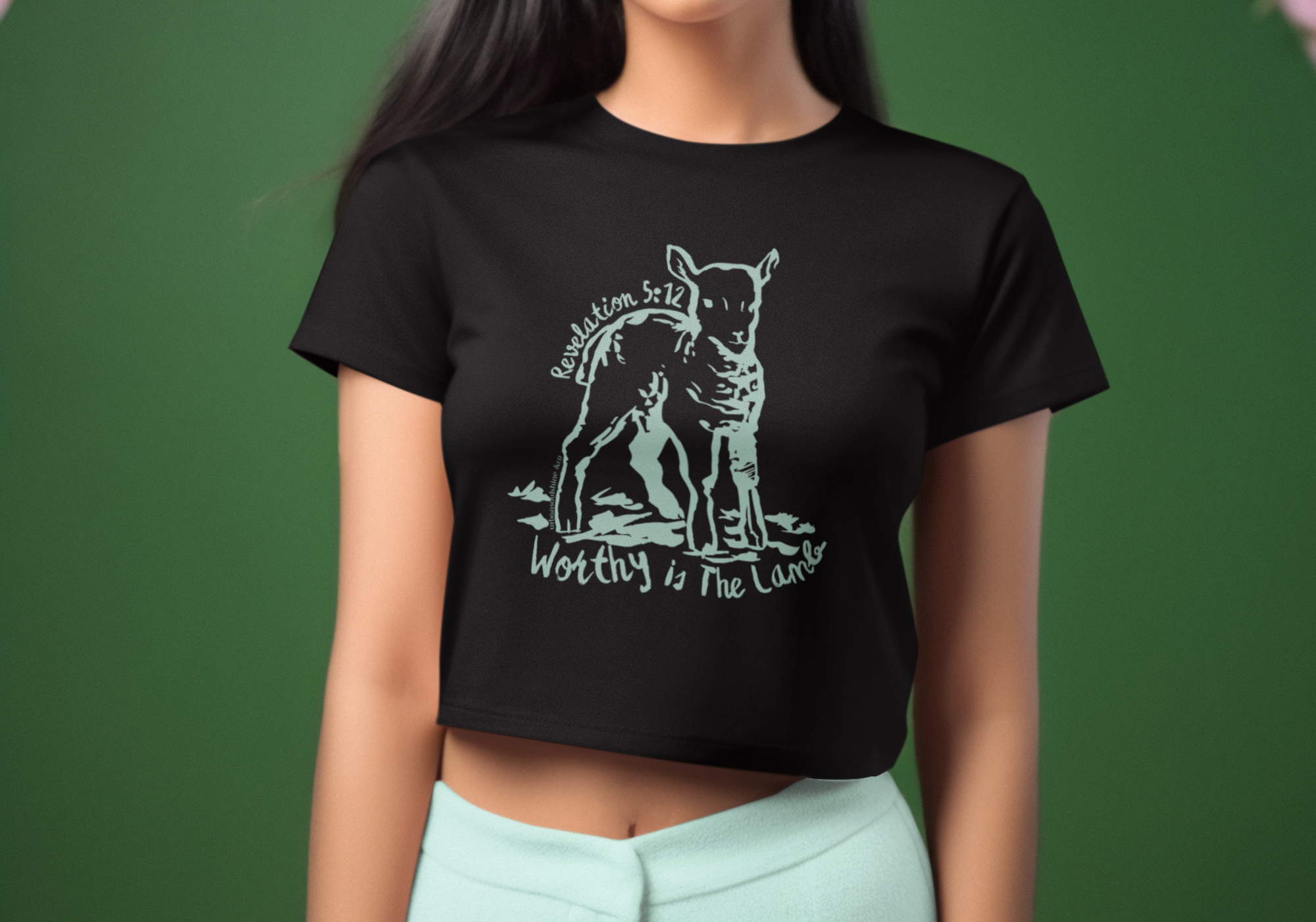 A woman stands in front of a green and pink backdrop wearing a black crop top with a dog illustration and text "Worthy is the Lamb" and "Revelation 5:12". She is also wearing light-colored pants and has straight dark hair.