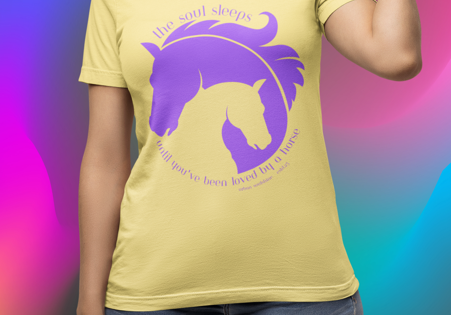 A person is wearing a yellow t-shirt featuring a purple graphic of a horse's head nestled with its foal's head. Above, text reads "the soul sleeps," and below, "until you've been loved by a horse." The background is a vibrant gradient of rainbow colors.