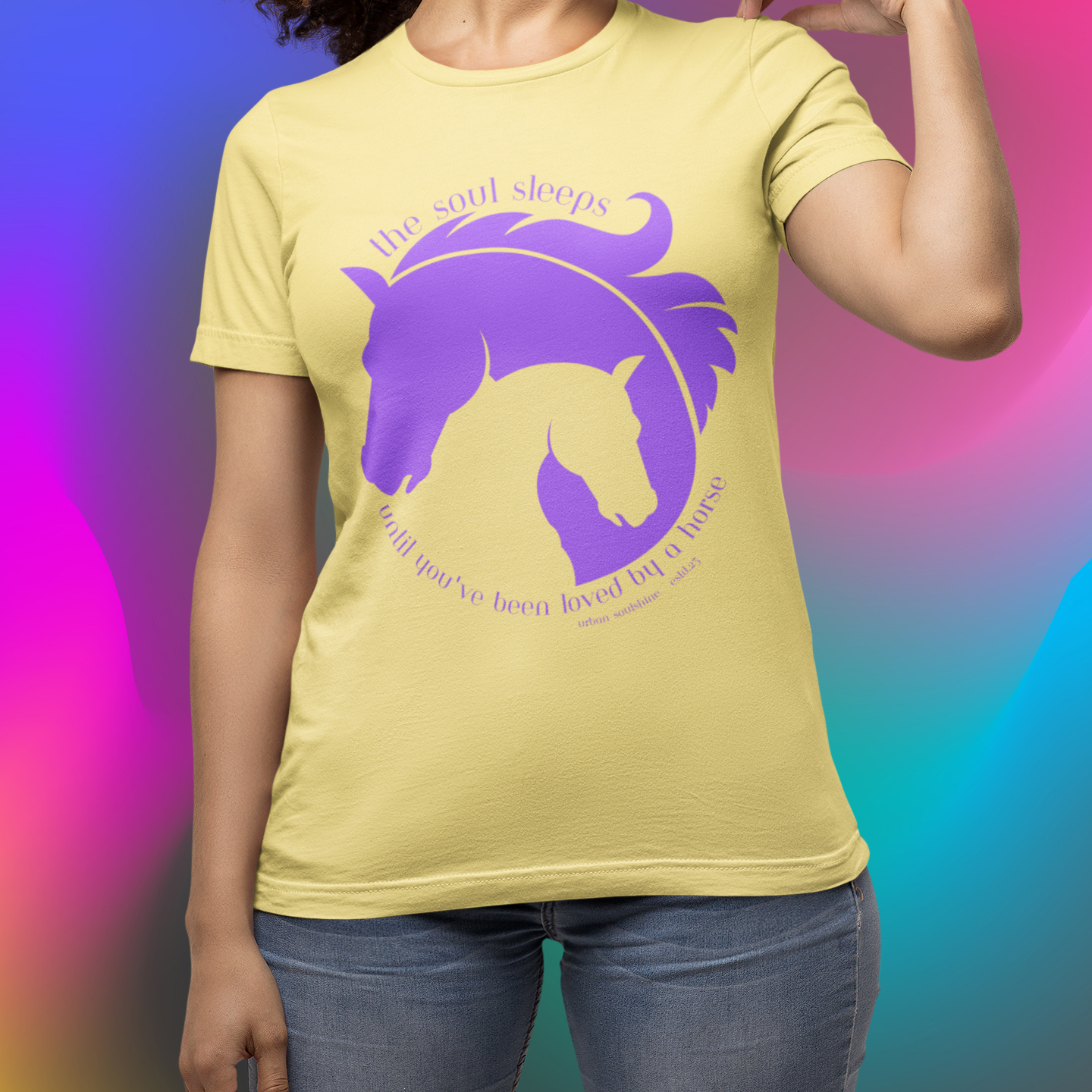 A person is wearing a yellow t-shirt featuring a purple graphic of a horse's head nestled with its foal's head. Above, text reads "the soul sleeps," and below, "until you've been loved by a horse." The background is a vibrant gradient of rainbow colors.