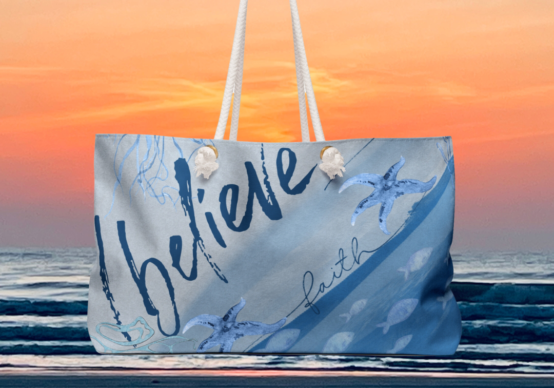 A beach bag with white handles is displayed against a backdrop of a sunset over the ocean. The bag features a blue and white design with starfish and the words "believe" and "faith" printed on it.