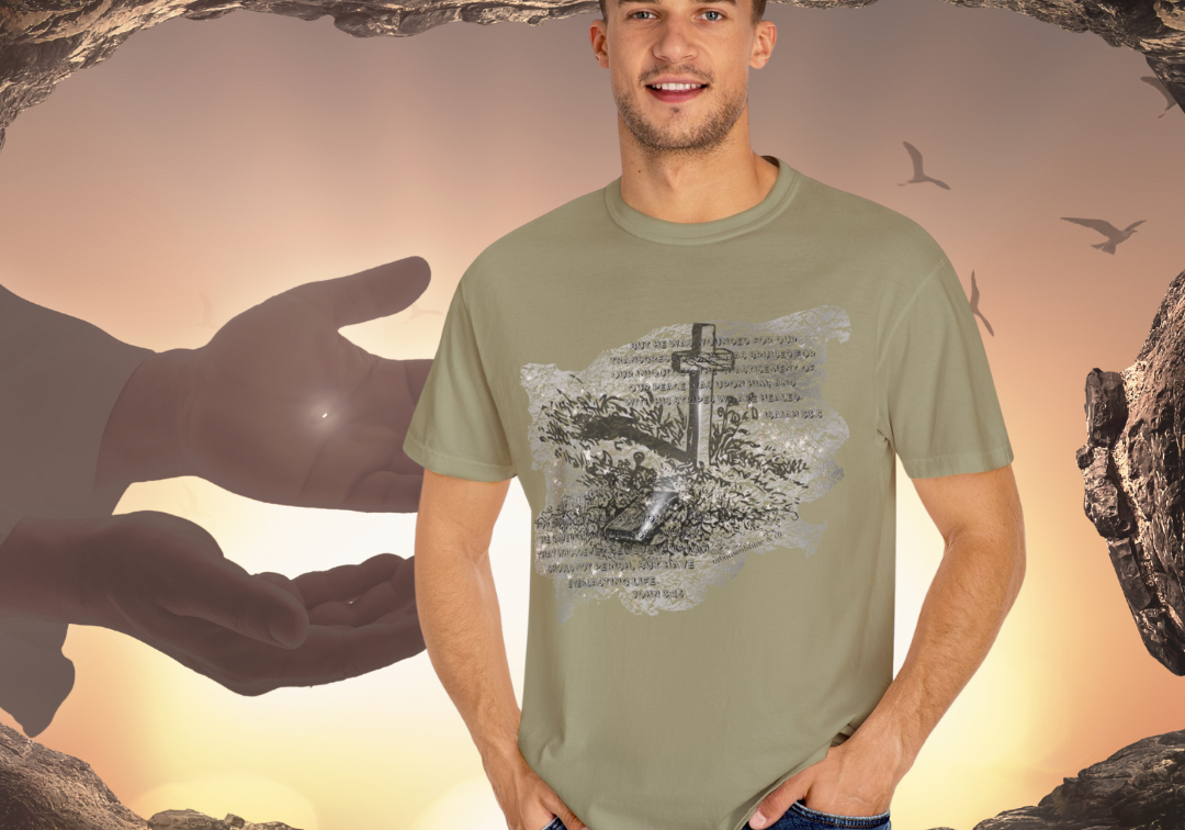 A man stands smiling, wearing a beige t-shirt with a graphic of a cross, rocks, and text. Background shows a sunrise or sunset with silhouetted birds and hands reaching out from a cave.