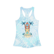 A person with long hair is wearing the Surf's Up Good Vibes Only Tie Dye Racerback Tank Top, made from lightweight fabric and featuring a graphic of a surfboard and palm trees with the message "Good Vibes Only" on the front. They are also dressed in light blue jeans from Urban SoulShine & co and are smiling at the camera.