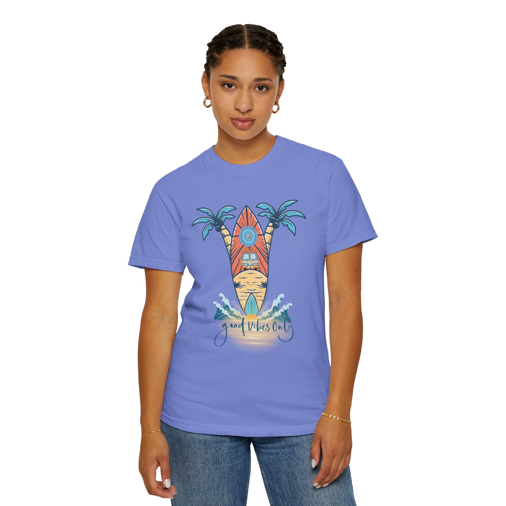 A person with short, braided hair is wearing a blue t-shirt with a design of two palm trees, an ocean wave, and a surfboard in between. The text on the shirt reads "Good Vibes Only." The person is also wearing hoop earrings and a bracelet that lets them express themselves. They are sporting the Surf's Up! Good Vibes Only Tee (Unisex) by Printify.