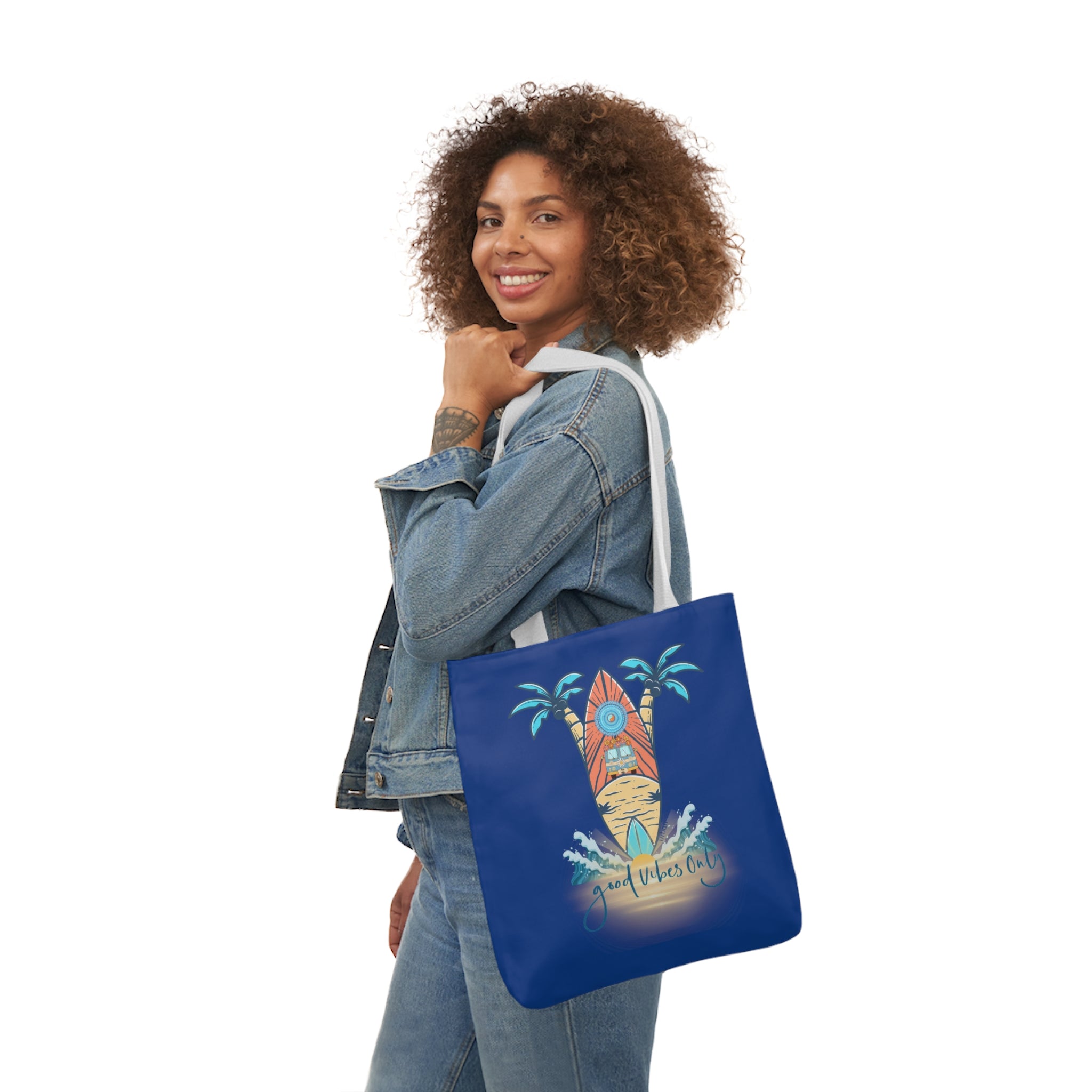 A person with curly hair smiles while holding a Surf's Up Good Vibes Only Beach Tote from Printify on their shoulder. The bag has a beach-themed design with a surfboard, palm trees, waves, and the text "Just takes it all." The person is wearing a denim jacket and blue jeans, standing against a white background.