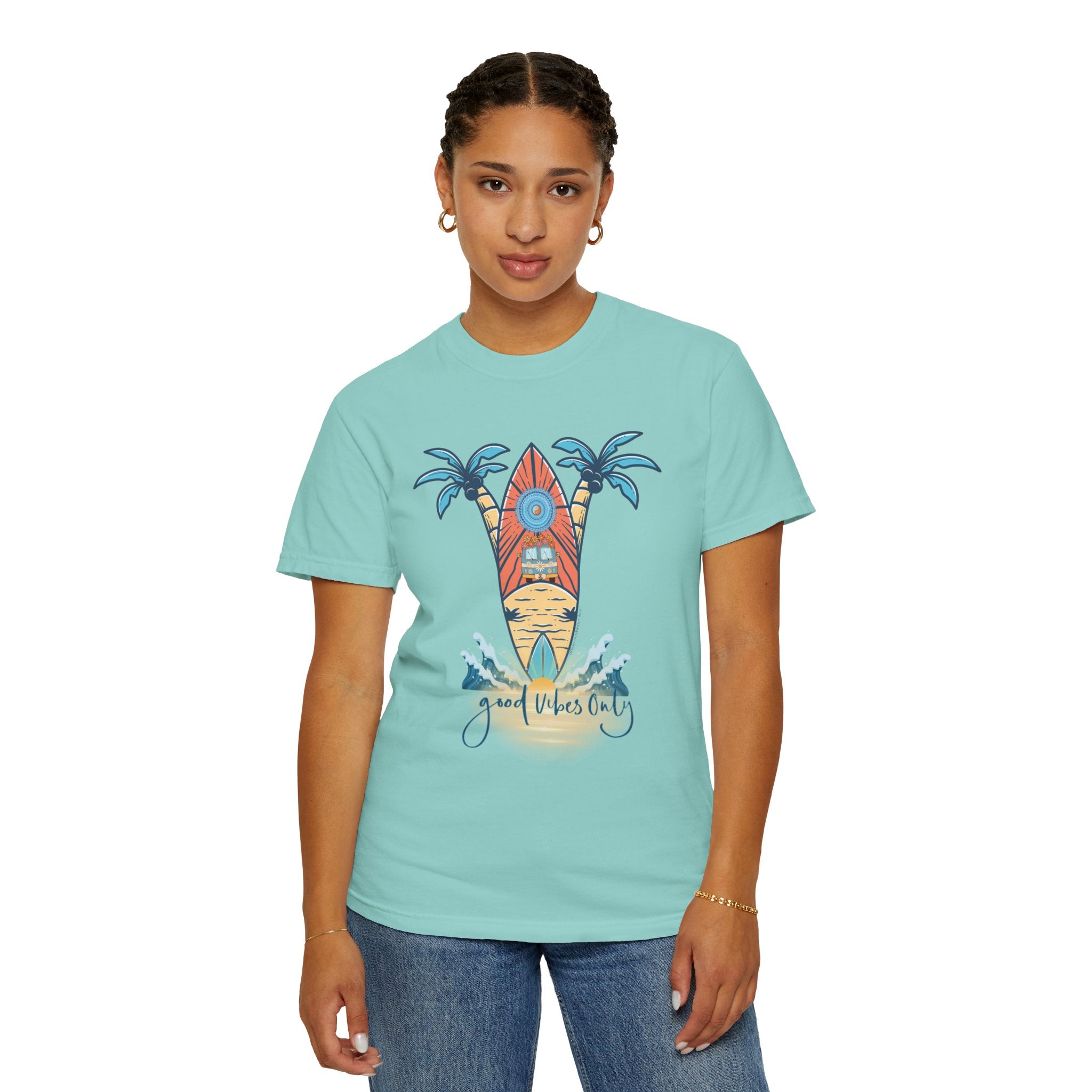 A person with short braids is standing and wearing a light blue Surf's Up! Good Vibes Only Tee (Unisex) by Printify featuring a colorful surfboard design with palm trees and the text "Good Vibes Only." They are also wearing jeans and simple jewelry, including hoop earrings and a bracelet. Their outfit truly reflects "soulshine.