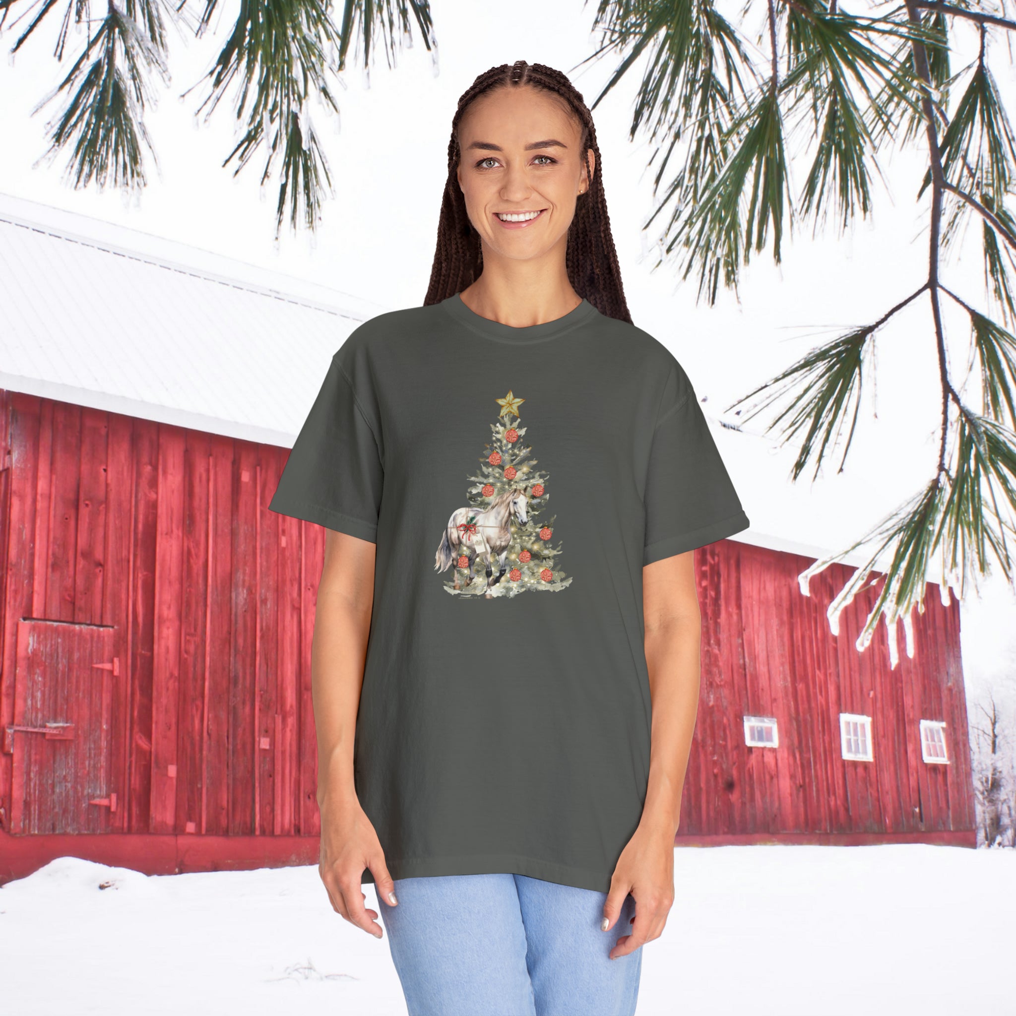 A person stands outdoors in front of a red barn with snow covering the ground, wearing The Gift of A Horse Christmas Garment-Dyed T-shirt by Urban SoulShine & co, which features a festive Christmas tree design adorned with animals and decorations. Pine branches can be seen above, while the cozy ring-spun cotton enhances the warmth of the wintry scene.