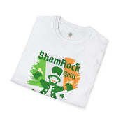 A man with curly hair and a beard sits in front of a bar counter, smiling. He's wearing a grey T-shirt with a colorful graphic of a leprechaun and text that reads "ShamRock Grill" and "McFizzle's Irish PUB" alongside a shamrock and musical notes. His Printify Shamrock Grill & McFizzie's Irish Pub SoftStyle Tee (Unisex) radiates soulshine, embodying the spirit of inspirational wear.