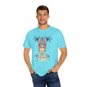 A man in a light blue T-shirt with a surfboard and palm tree design stands against a plain white background. The text on the T-shirt reads "Good Vibes Only." He has short brown hair and is wearing blue jeans, hands in his pockets, channeling artistic expression. The T-shirt he's wearing is the Surf's Up! Good Vibes Only Tee (Unisex) by Printify.