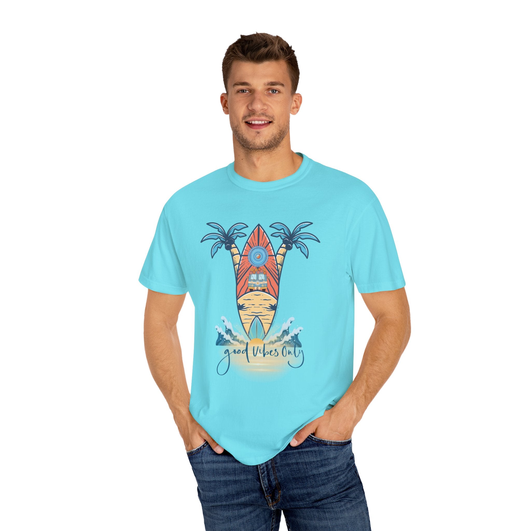 A man in a light blue T-shirt with a surfboard and palm tree design stands against a plain white background. The text on the T-shirt reads "Good Vibes Only." He has short brown hair and is wearing blue jeans, hands in his pockets, channeling artistic expression. The T-shirt he's wearing is the Surf's Up! Good Vibes Only Tee (Unisex) by Printify.