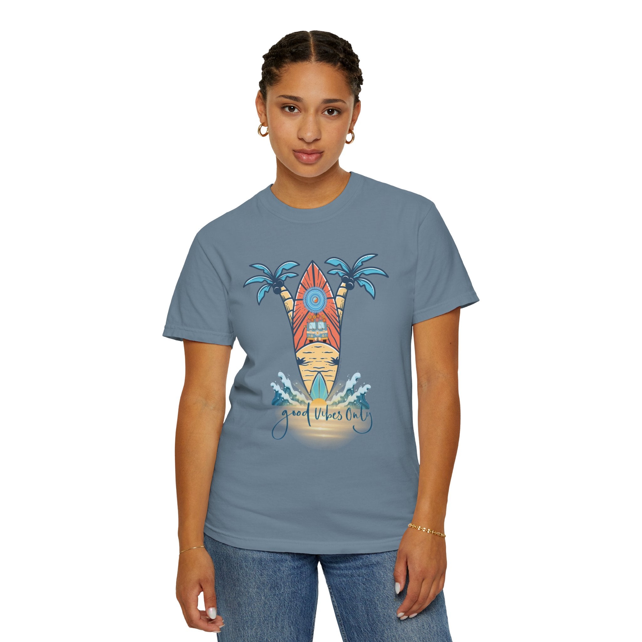 A person wearing a blue T-shirt with a colorful graphic of a surfboard, palm trees, and the text "Good Vibes Only." The person has short braided hair and is standing against a plain white background. They are also wearing jeans and gold hoop earrings, embodying artistic expression in their Surf's Up! Good Vibes Only Tee (Unisex) by Printify.