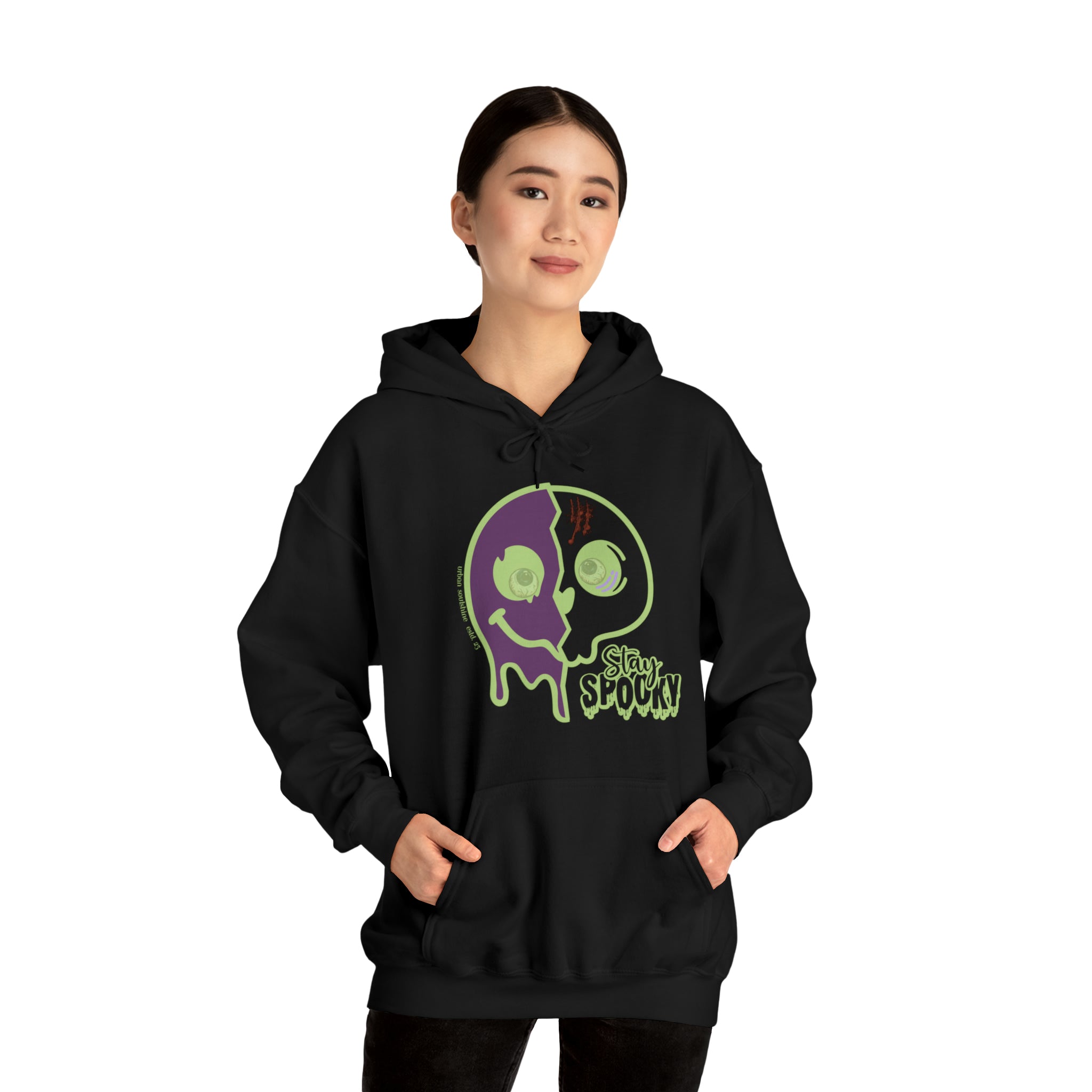 A person with tied-back hair is wearing a black hoodie featuring a green and purple skull design with the text "Stay Spooky" on the front. They have their hands in the hoodie pockets and are standing against a plain white background, showcasing their artistic expression in the Stay Spooky Funky Faces Halloween Hoodie (unisex) by Printify.