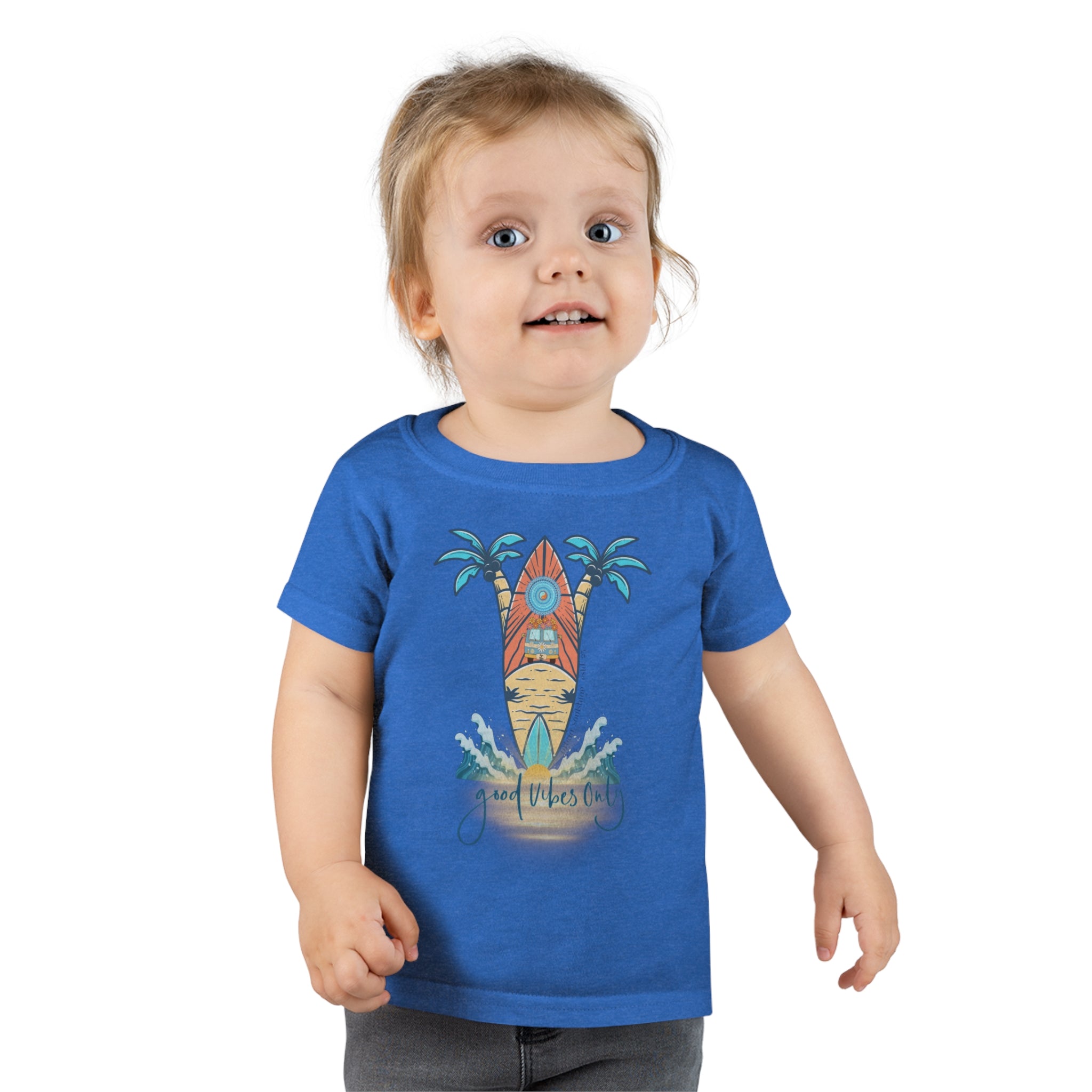 A smiling toddler with light brown hair is wearing a Surf's Up Good Vibes Only Toddler T-shirt by Printify featuring a colorful surfboard design with palm trees and waves. The text "good vibes only" is printed below the surfboard, capturing the spirit of inspirational wear. The child stands against a plain background.