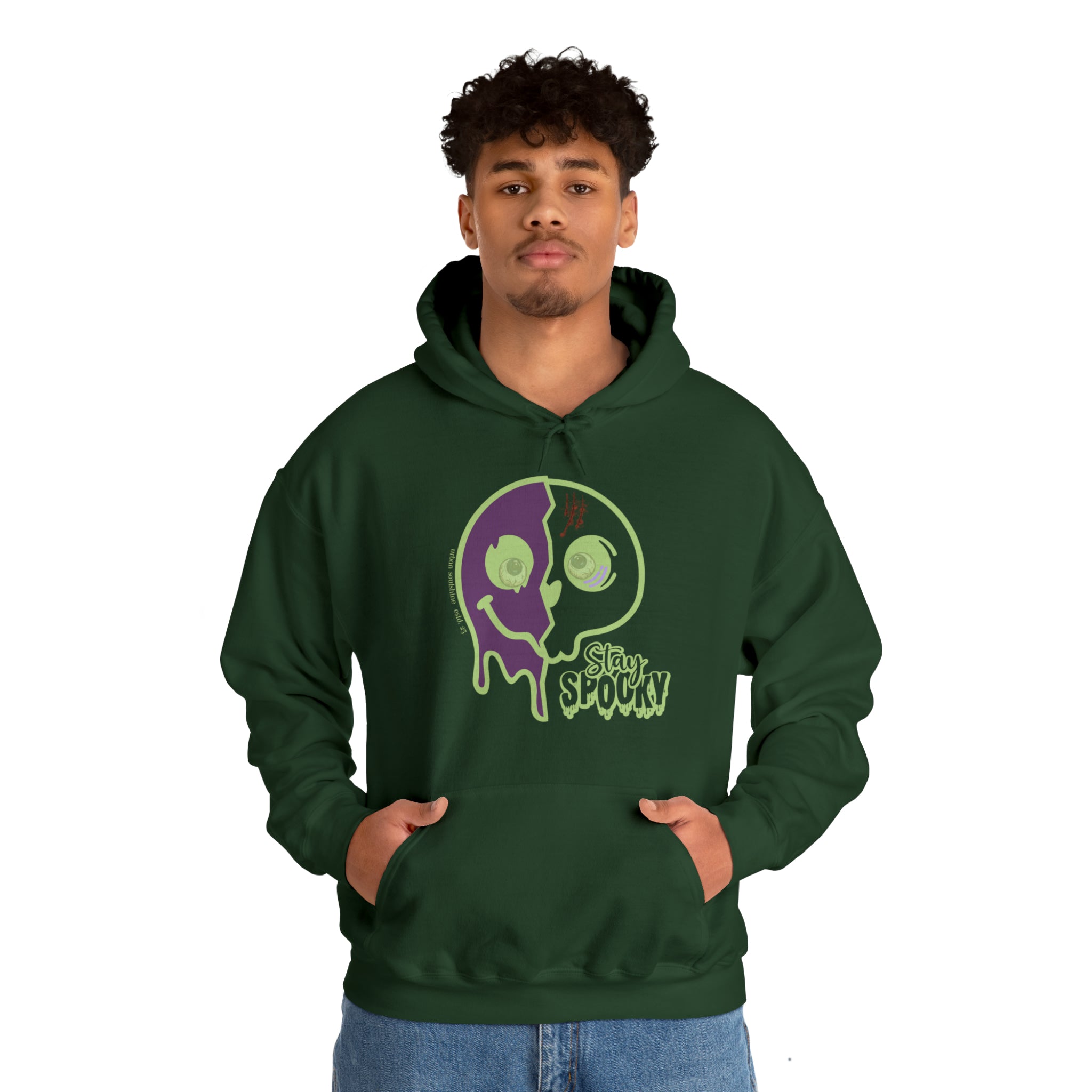 A man with curly hair is wearing a dark green Stay Spooky Funky Faces Halloween Hoodie (unisex) by Printify featuring a graphic design of a dripping, two-colored smiley face and the text "Stay Spooky." Expressing himself confidently, he stands with his hands in the front pocket of the hoodie and wears light blue jeans.