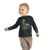 A young child with a slight smile stands against a plain background, wearing a gray long-sleeve shirt and blue jeans. The Spooky Island Halloween Toddler Long Sleeve Tee by Printify features a green, pumpkin-headed figure, a haunted house, and the phrase "Trick or Treat," showcasing an artistic expression perfect for the season.