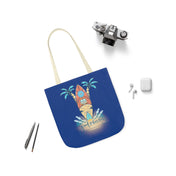 A lightweight blue polyester canvas tote bag with comfortable tan handles is decorated with an illustration of a tiki statue, palm trees, ocean waves, and the phrase "Good Vibes Only." Surrounding the Urban SoulShine & co Surf's Up Good Vibes Only Beach Tote are a vintage camera, two pens, wireless earbuds, and an AirPods case.