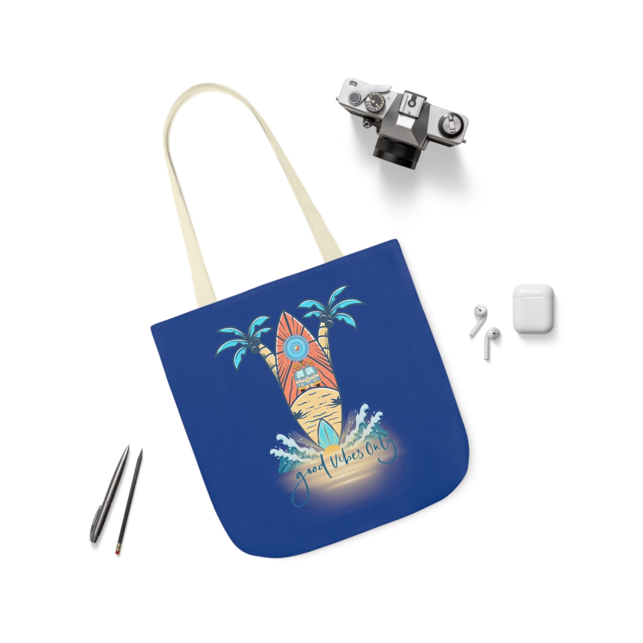 A blue tote bag with a design featuring a surfboard, palm trees, and the text "Good Vibes Only" is surrounded by a camera, earphones, an AirPods case, and two pens on a white background. Express yourself with the Surf's Up Good Vibes Only Beach Tote from Printify.