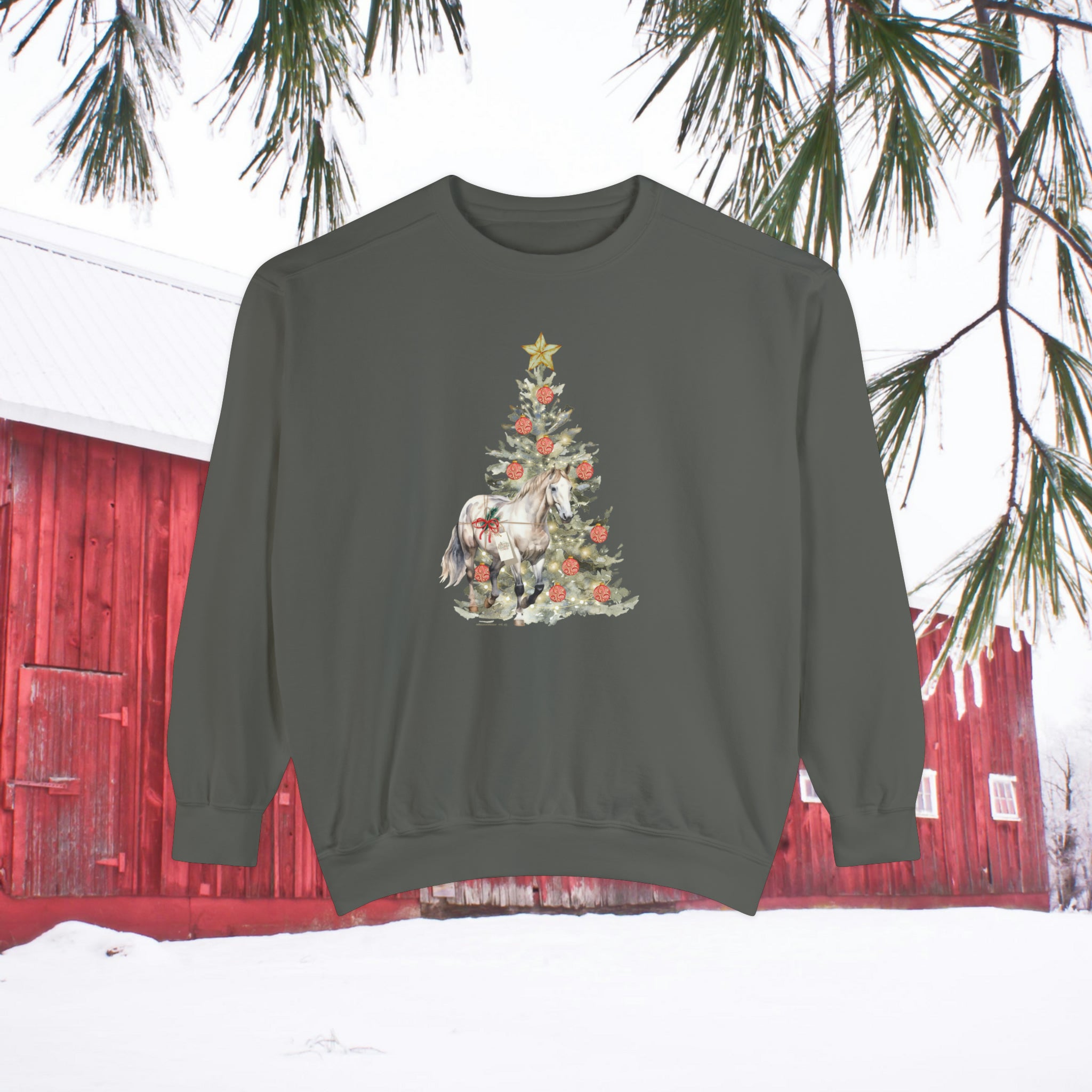 The Gift of A Horse Christmas Garment-Dyed Sweatshirt by Urban SoulShine & co is showcased outdoors against a backdrop of a red barn and snowy terrain. This unisex sweatshirt, with a relaxed fit and dark green color, highlights a Christmas tree topped with a star and adorned with festive decorations, along with a horse at its base. Overhead, pine branches add to the seasonal ambiance.