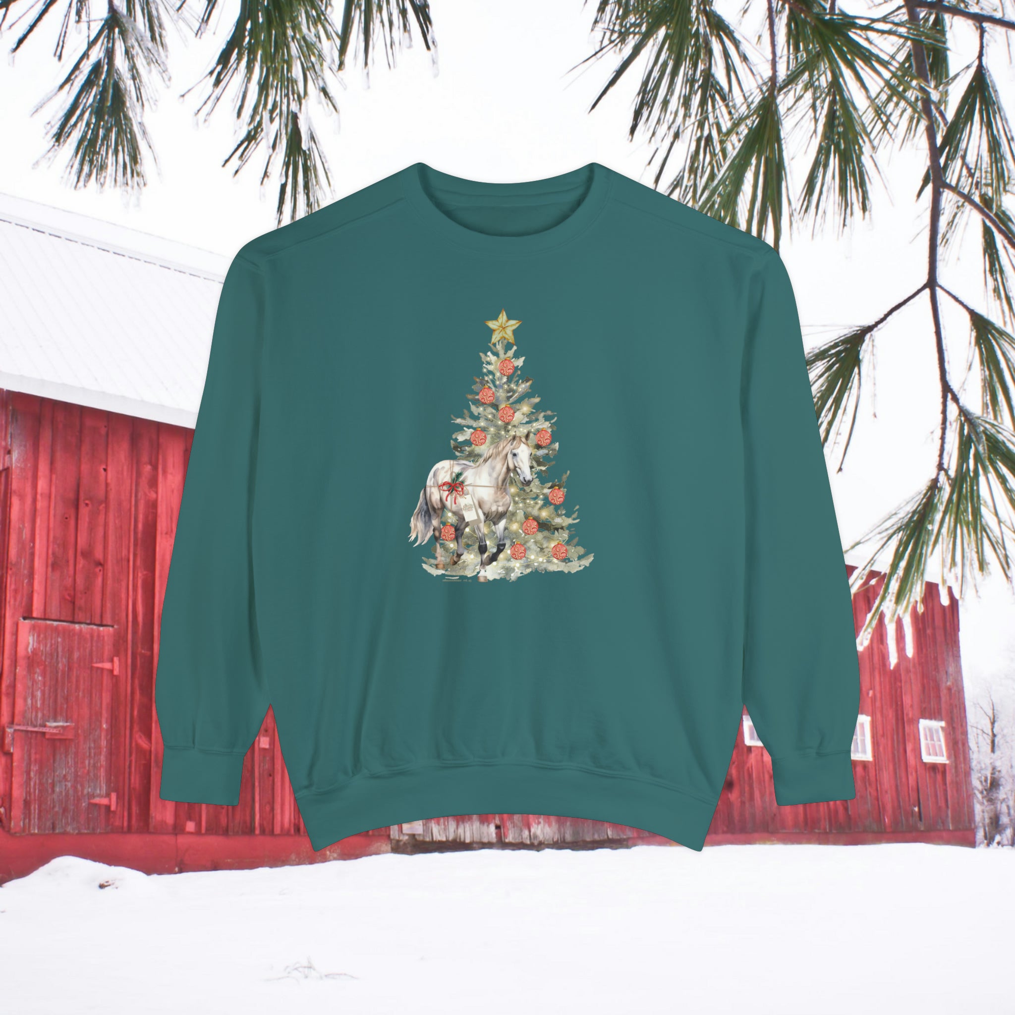 Displayed outdoors, "The Gift of A Horse Christmas Garment-Dyed Sweatshirt" by Urban SoulShine & co features a relaxed fit in a green unisex design adorned with a unicorn and Christmas tree. The cozy holiday scene includes a red barn backdrop and a snow-covered landscape decorated with pine branches.