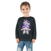 A young girl smiles at the camera, wearing a black Printify Too Cute To Spook Halloween Toddler Long Sleeve Tee (unisex Tot) featuring cartoon ghost faces and the words "Too Cute to Spook." She also wears light-wash, ripped jeans and has her hair styled in a ponytail. Her outfit is perfect for those who love to express yourself.