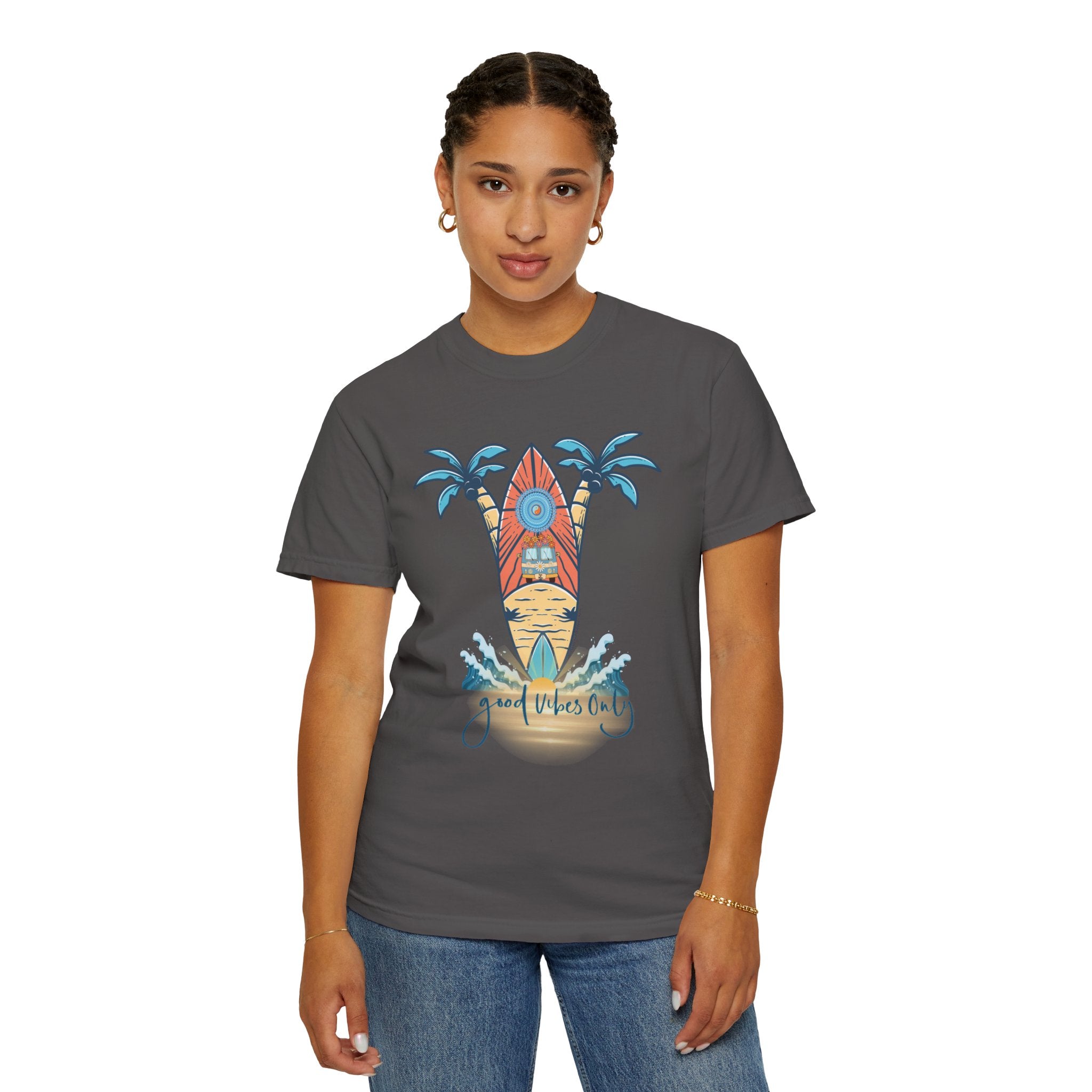 A person stands facing the camera wearing a charcoal gray Surf's Up! Good Vibes Only Tee (Unisex) by Printify with a colorful surfboard and palm tree design, along with the text "Good Vibes Only" on the front. The t-shirt captures an artistic expression that resonates. The person is also wearing jeans and has short braided hair.