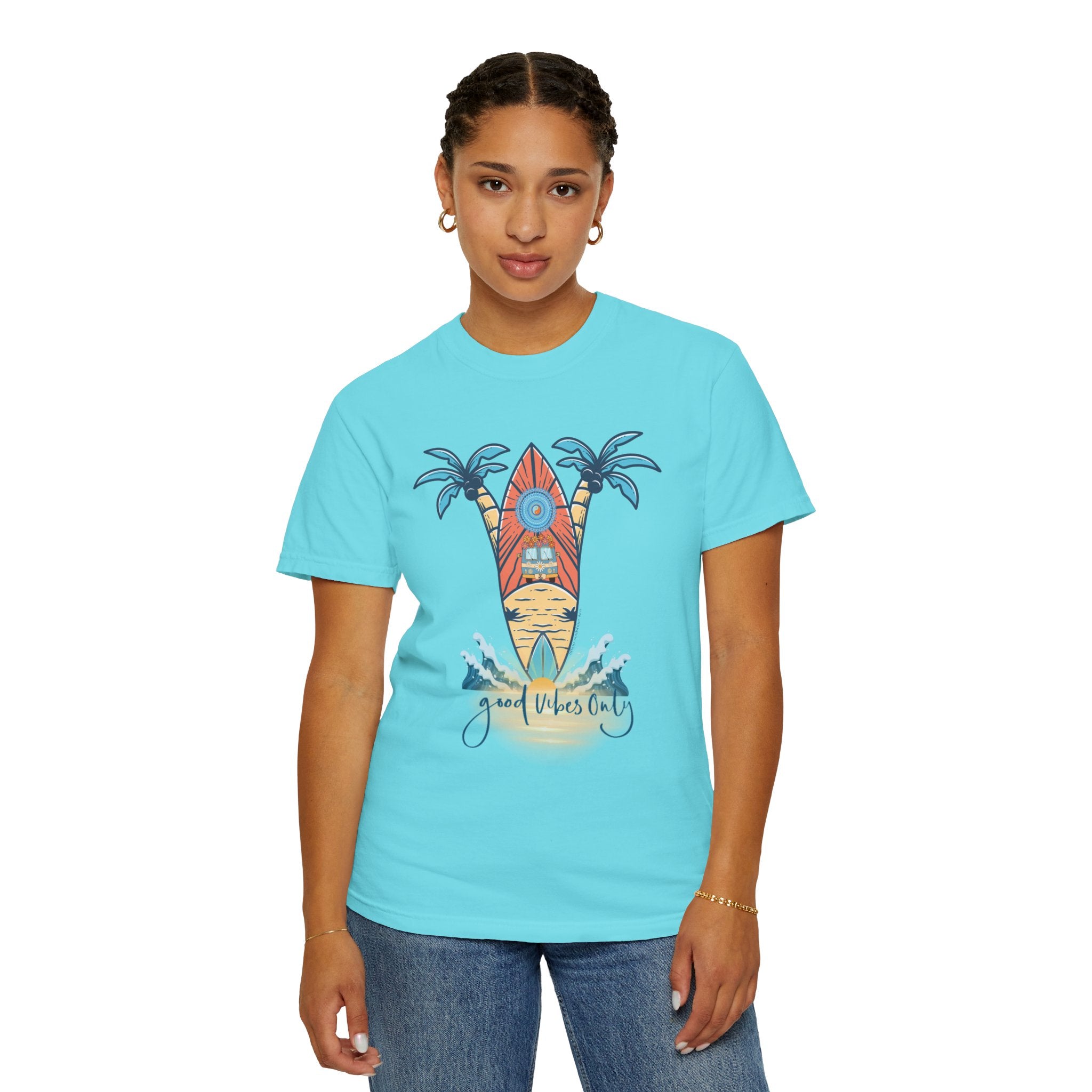 A person with braided hair, wearing a turquoise t-shirt featuring a surfboard, palm trees, and the words "Good Vibes Only," stands against a plain white background. This piece of inspirational wear, the Surf's Up! Good Vibes Only Tee (Unisex) by Printify, is paired with blue jeans and small hoop earrings.