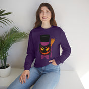 A person with long brown hair is smiling and sitting on a white surface. They are wearing an Urban SoulShine & co Top Hat Jack Halloween Crewneck Sweatshirt (unisex) with a Halloween-themed graphic of a pumpkin, black top hat, and broomstick. A plant in a white pot is to the left.