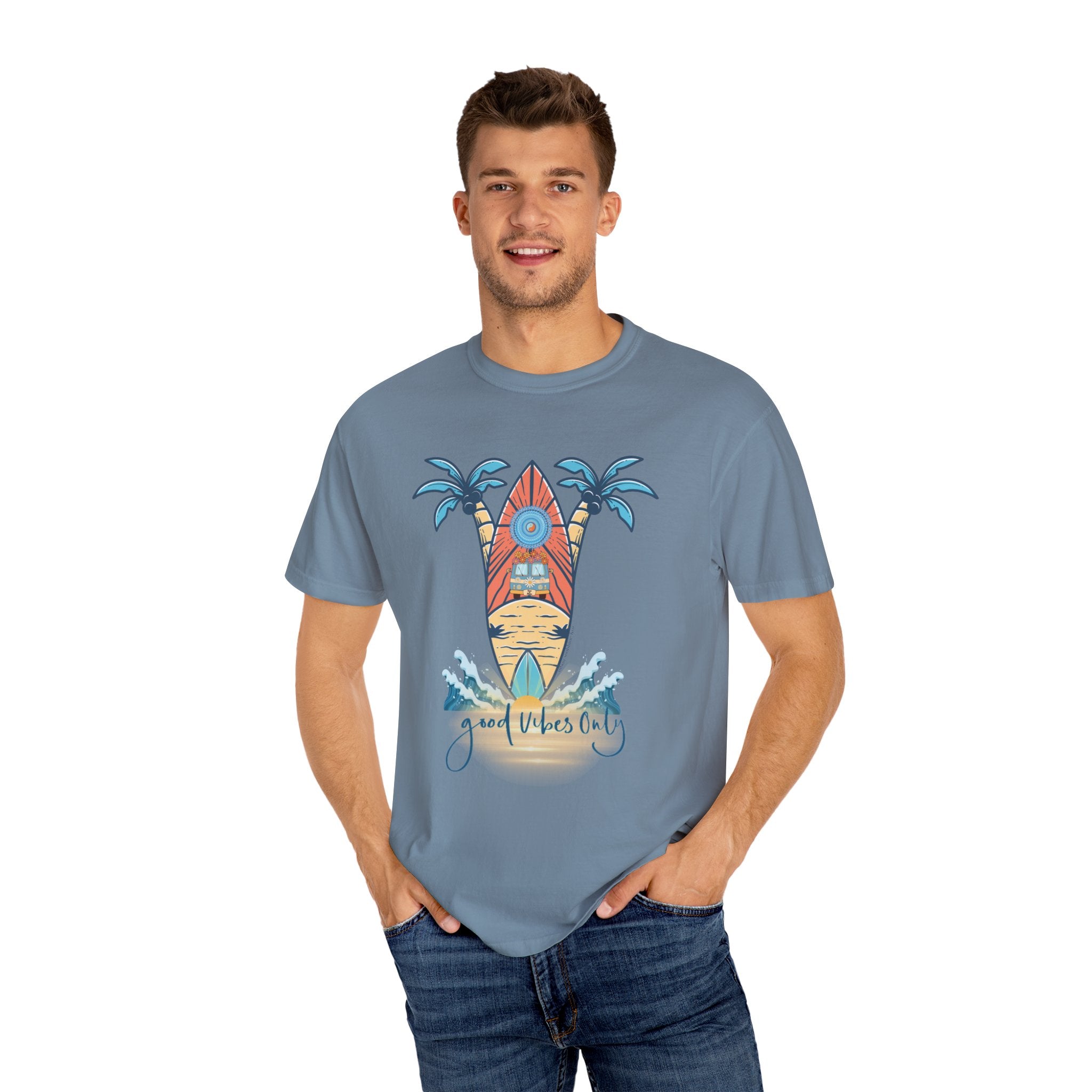 A man stands smiling, wearing a light blue t-shirt with a surfboard and palm tree graphic. The text on the shirt reads "Good Vibes Only." He has his hands in the pockets of his blue jeans and is set against a plain white background, showcasing the Surf's Up! Good Vibes Only Tee (Unisex) by Printify to express yourself.