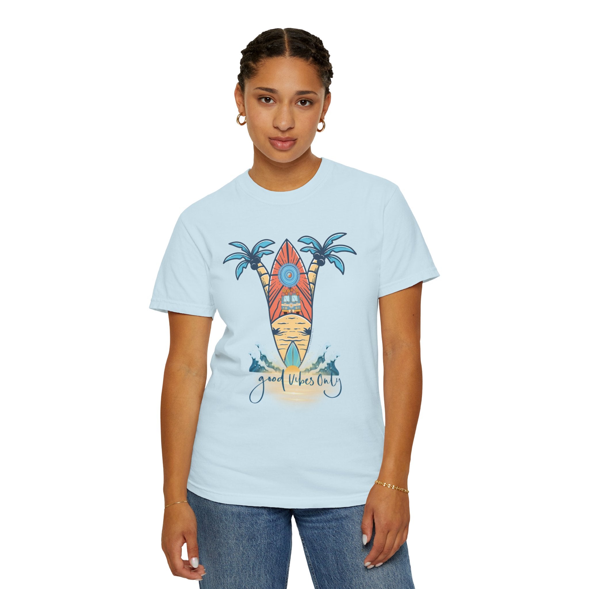 A person wearing the Surf's Up! Good Vibes Only Tee (Unisex) by Printify, which features a graphic of two surfboards and palm trees with the text "Good Vibes Only." The individual has short braided hair, small hoop earrings, a bracelet, and is standing against a plain white background. Dare to inspire with your unique style and artistic expression.