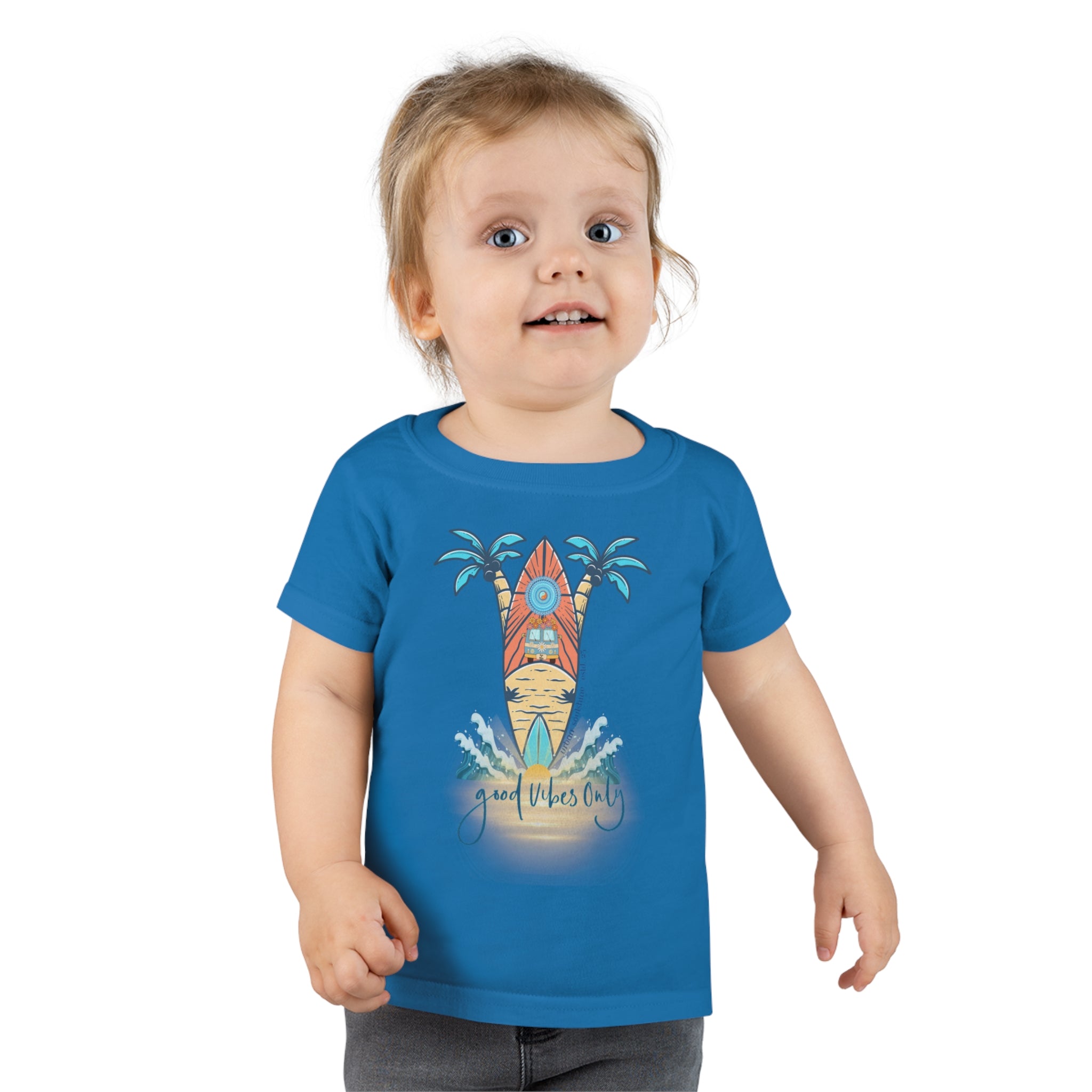 A smiling toddler with light brown hair is wearing a Surf's Up Good Vibes Only Toddler T-shirt by Printify featuring a colorful surfboard design with palm trees and waves. The text "good vibes only" is printed below the surfboard, capturing the spirit of inspirational wear. The child stands against a plain background.