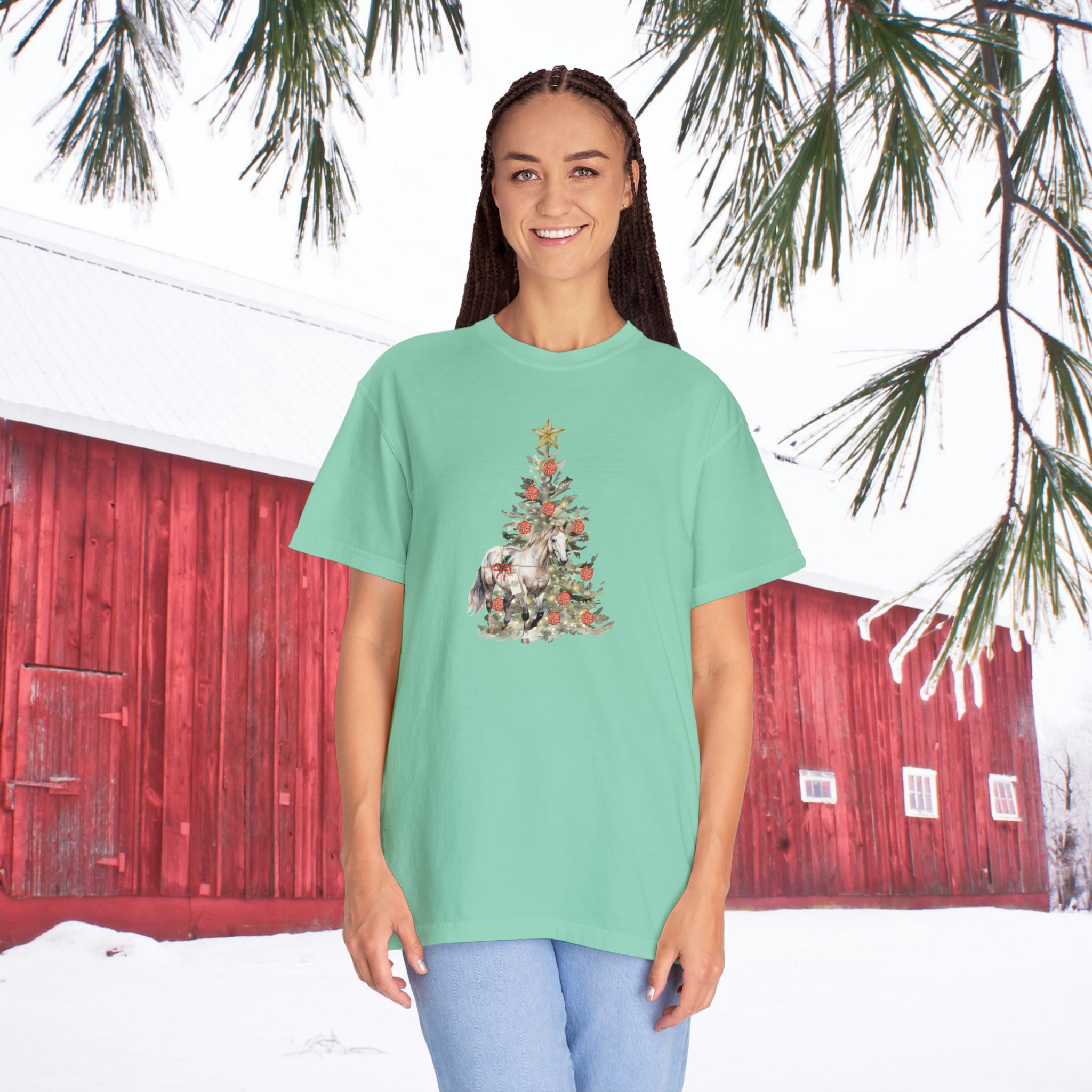 A person stands outside in the snow wearing The Gift of A Horse Christmas Garment-Dyed T-shirt by Urban SoulShine & co. A red barn and snowy pine branches provide a picturesque backdrop, highlighting the cozy ring-spun cotton of their outfit.