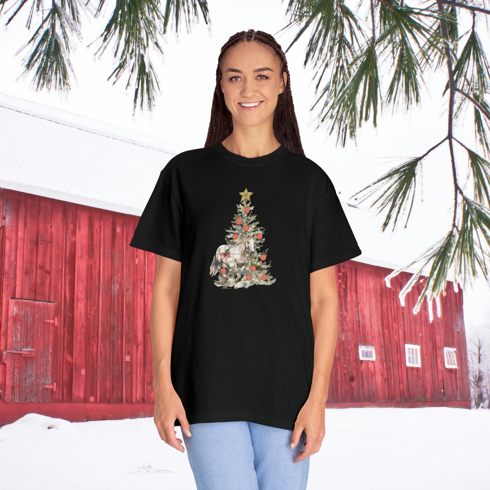 A person wearing "The Gift of A Horse Christmas Garment-Dyed T-shirt" by Urban SoulShine & Co. stands outside. The black t-shirt showcases a Christmas tree design, creating a striking contrast against the snow-covered ground, with a red barn and pine tree branch in the background.