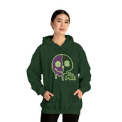 A person with dark hair is standing and wearing a dark green hoodie with a graphic design featuring a purple and green melting skull and the text "Stay Spooky." They are facing the camera, hands in their pockets. This Printify Stay Spooky Funky Faces Halloween Hoodie (unisex) invites you to embrace your unique style and dare to inspire.