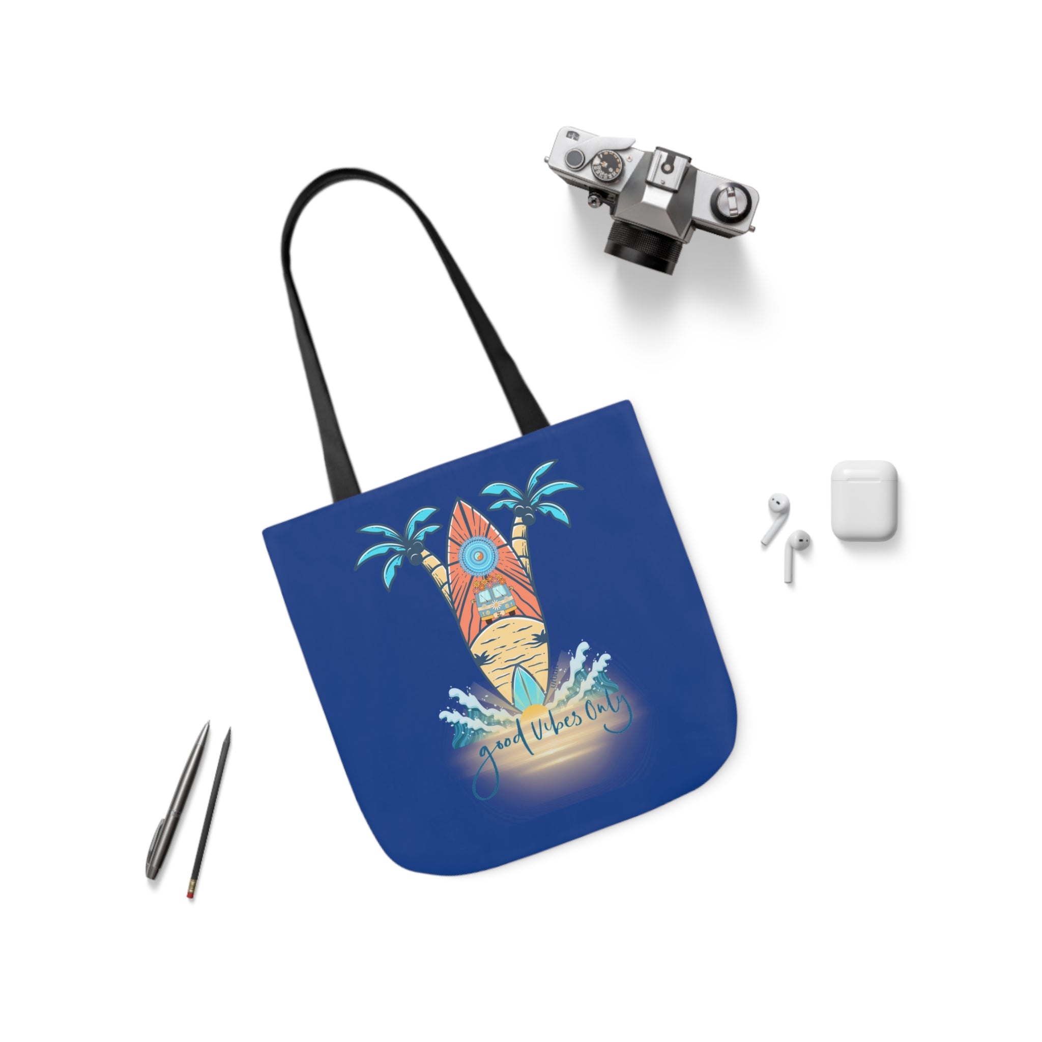 A blue Printify Surf's Up Good Vibes Only Beach Tote with black straps features an illustration of a surfboard between two palm trees and the words "Good Vibes Only" written below in a beach-themed design. Surrounding the tote are a vintage camera, a pair of earbuds, a white case, and two pens. Dare to inspire everywhere you go!