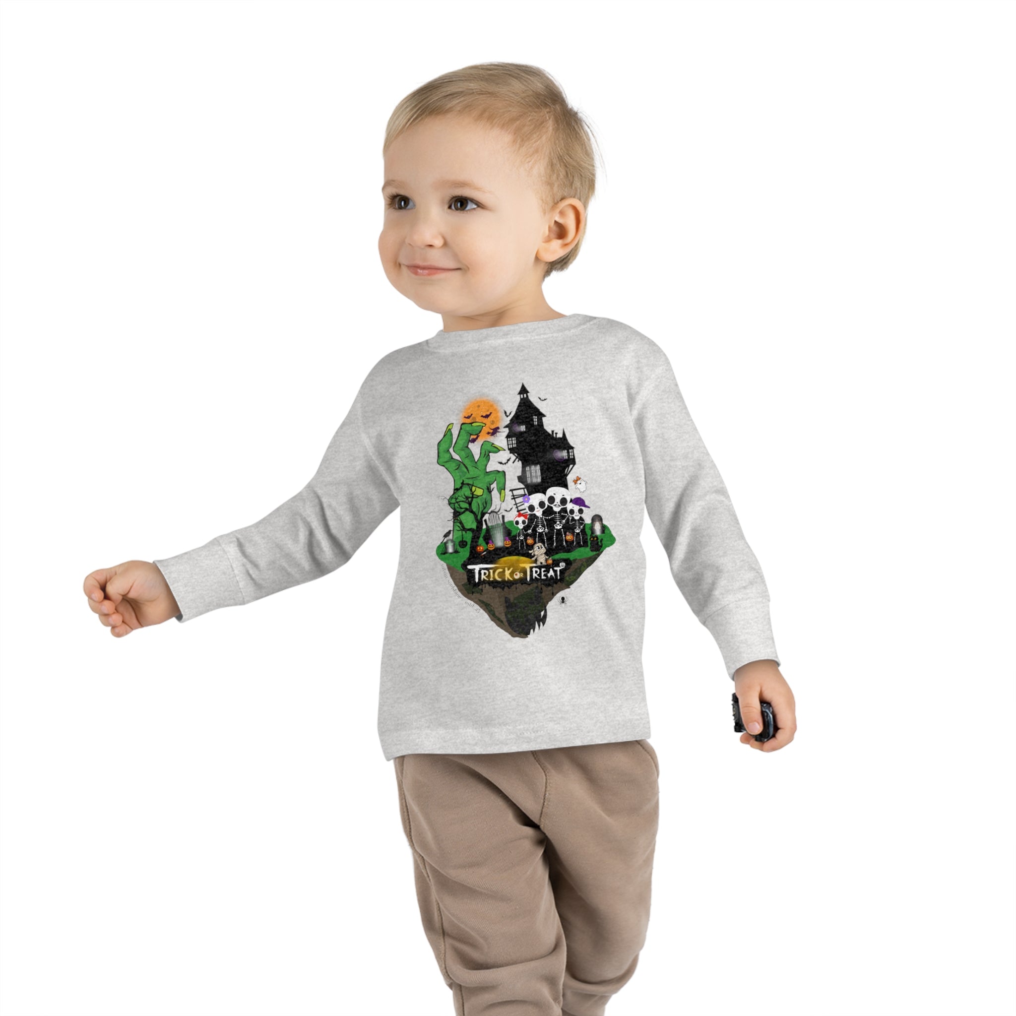 A young girl with a ponytail is wearing the Spooky Island Halloween Toddler Long Sleeve Tee by Urban SoulShine & Co. This 100% combed ringspun cotton shirt features a Halloween-themed design with the text "Trick Or Treat" and illustrated characters. She is smiling and standing with her hands by her sides, also sporting distressed jeans.