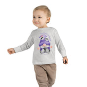 A young child with short blond hair is wearing a gray long-sleeve shirt and brown pants. The shirt features an illustration of two small Halloween creatures dressed in purple, with the words "Too Cute to Spook" above them. The child, embodying artistic expression, is smiling and holding a small object. They are wearing the Printify Too Cute To Spook Halloween Toddler Long Sleeve Tee (unisex Tot).