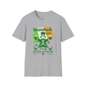 A man with curly hair and a beard sits in front of a bar counter, smiling. He's wearing a grey T-shirt with a colorful graphic of a leprechaun and text that reads "ShamRock Grill" and "McFizzle's Irish PUB" alongside a shamrock and musical notes. His Printify Shamrock Grill & McFizzie's Irish Pub SoftStyle Tee (Unisex) radiates soulshine, embodying the spirit of inspirational wear.