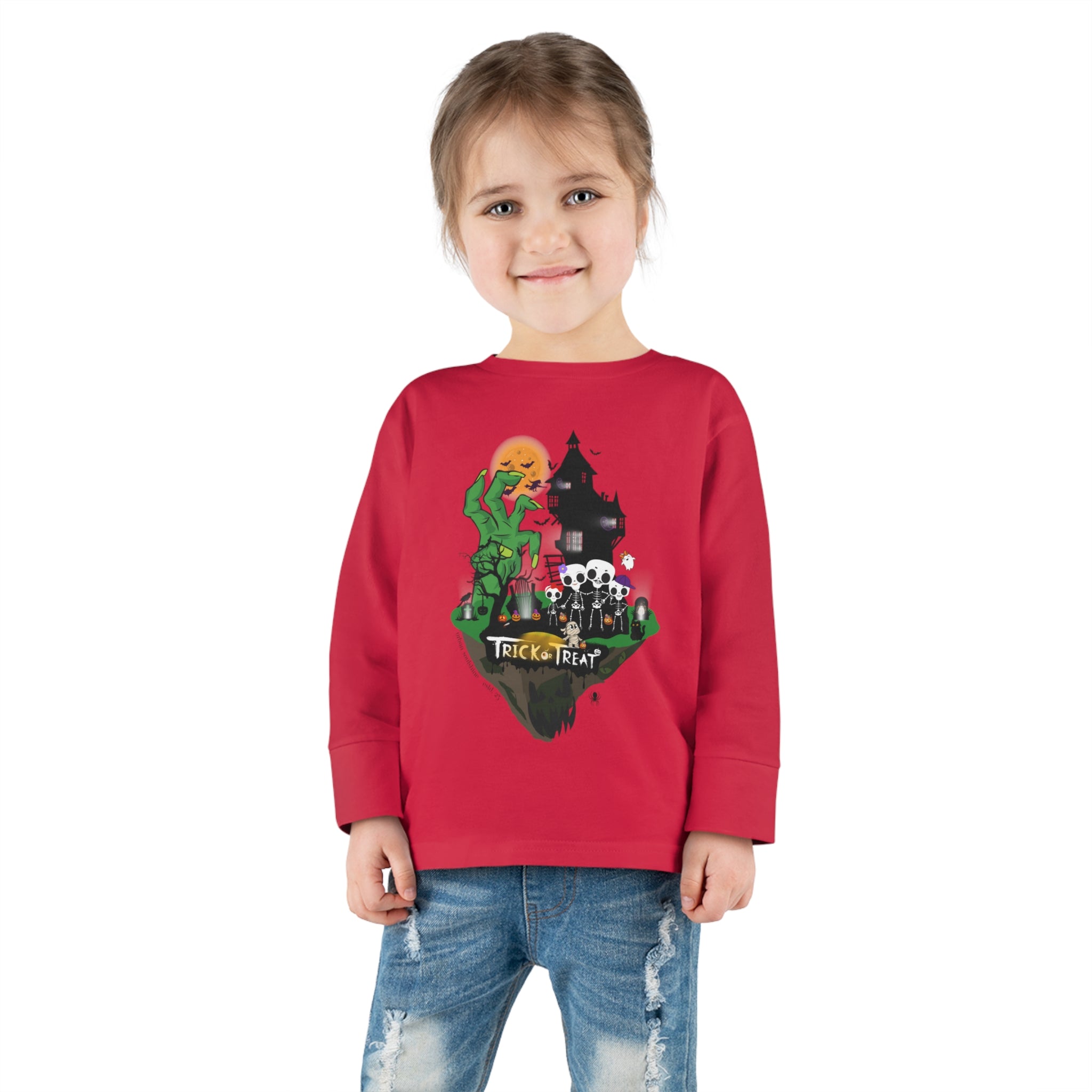 A young girl with a ponytail is wearing the Spooky Island Halloween Toddler Long Sleeve Tee by Urban SoulShine & Co. This 100% combed ringspun cotton shirt features a Halloween-themed design with the text "Trick Or Treat" and illustrated characters. She is smiling and standing with her hands by her sides, also sporting distressed jeans.