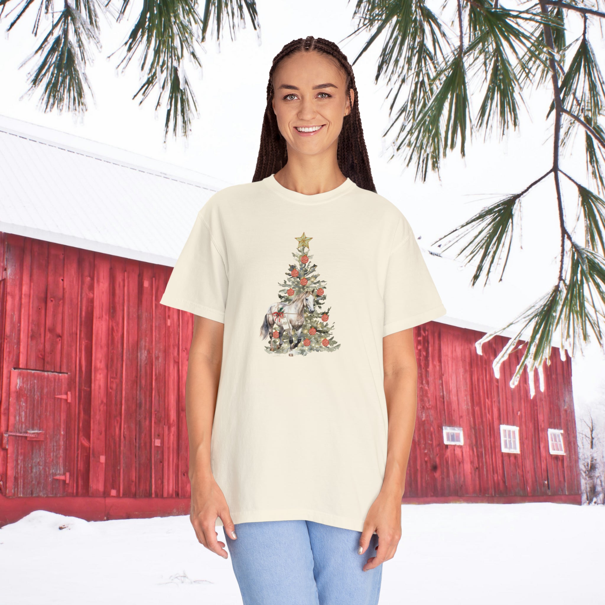 In a snowy landscape, with pine branches swaying gently overhead, a person smiles in front of a red barn while wearing The Gift of A Horse Christmas Garment-Dyed T-shirt by Urban SoulShine & Co. Adorned with charming Christmas tree and animal designs, the ring-spun cotton garment provides extra comfort and is perfectly paired with blue pants, adding to the cozy scene.