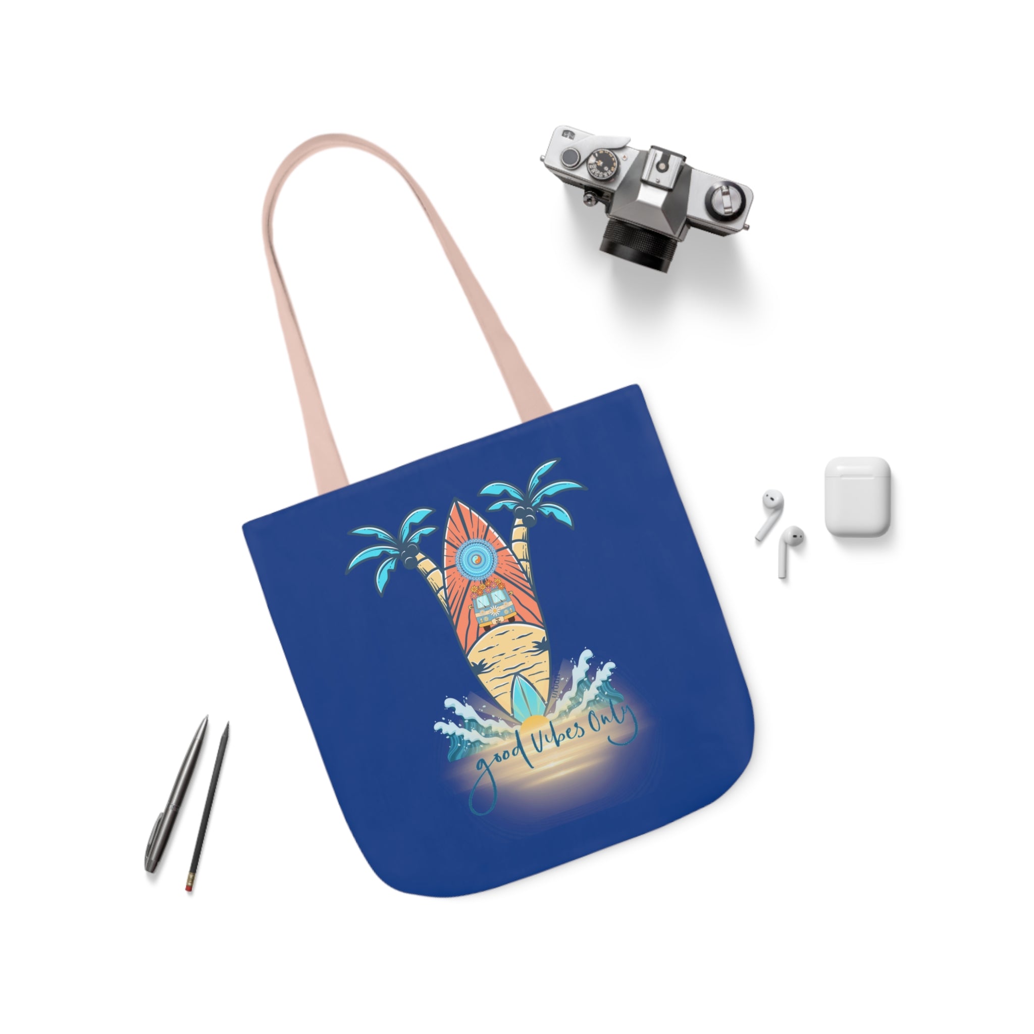 A blue tote bag with a design featuring an orange surfboard, palm trees, and the text "Good Vibes Only." The Printify Surf's Up Good Vibes Only Beach Tote is placed on a white surface alongside a vintage camera, wireless earbuds, and two pens. This stylish accessory embodies artistic expression and adds a touch of soulshine to your day.