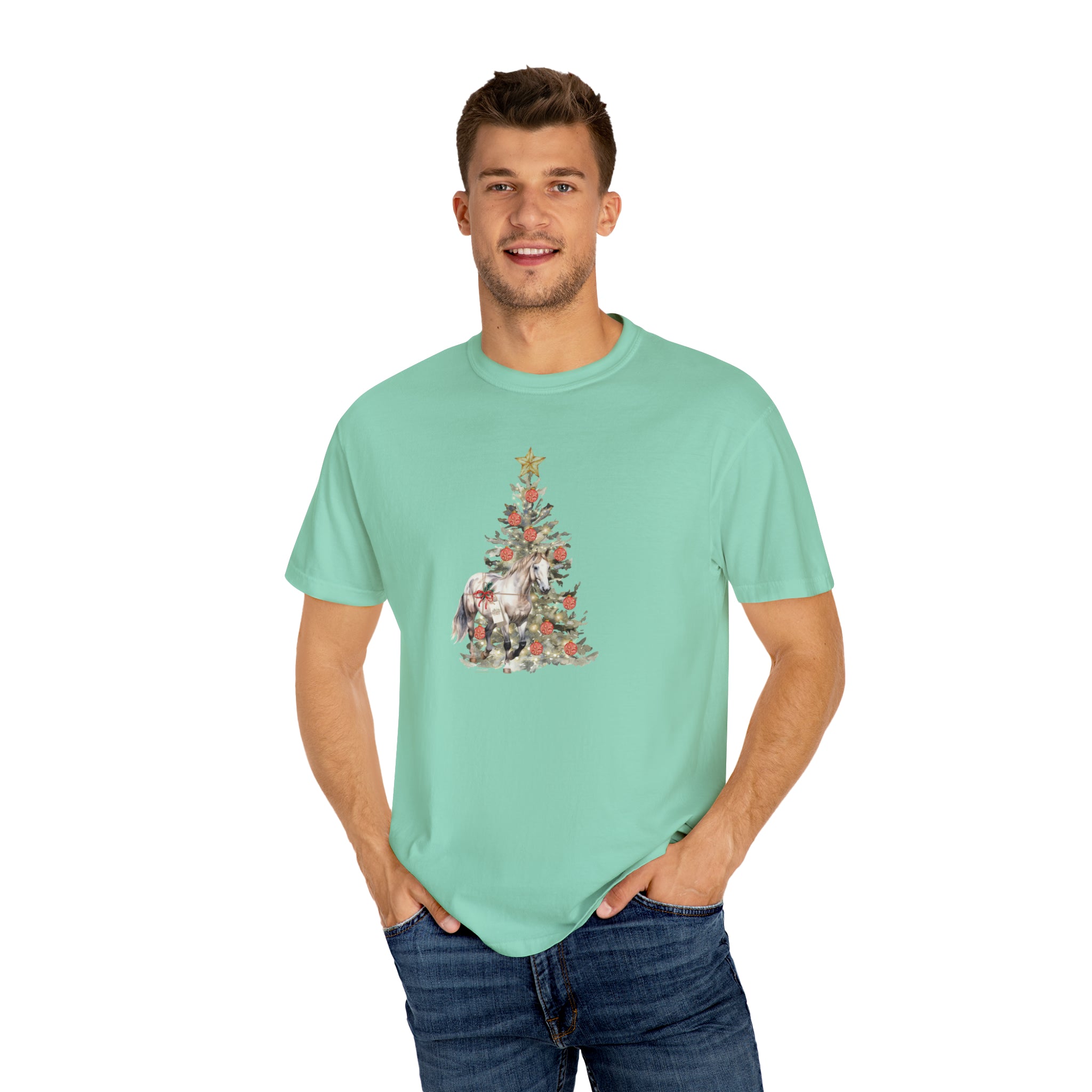 A man with short hair is wearing "The Gift of A Horse Christmas Garment-Dyed T-shirt" from Urban SoulShine & co, featuring a light green hue with a festive Christmas tree and decorated gifts on the front. He pairs this cheerful holiday design with blue jeans against a plain white background.