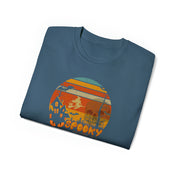 A folded blue Spooky Nights Halloween Ultra Cotton Tee (Unisex) by Printify with a Halloween-themed graphic in the center. The graphic features a witch flying on a broomstick over a sunset, with silhouettes of a graveyard, haunted house, and bats. The word "SPOOKY" is written at the bottom in orange font, inviting you to express yourself this Halloween.