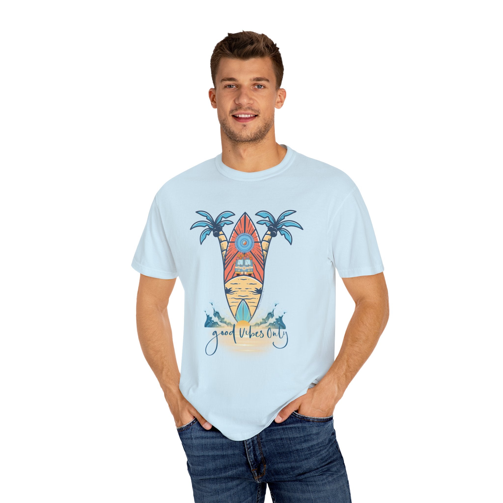 A man with short hair is wearing a Surf's Up! Good Vibes Only Tee (Unisex) by Printify featuring a surfboard, palm trees, and the text "Good Vibes Only" printed on the front. He is smiling, has his hands in his jeans pockets, and is standing against a plain white background exuding pure soulshine.