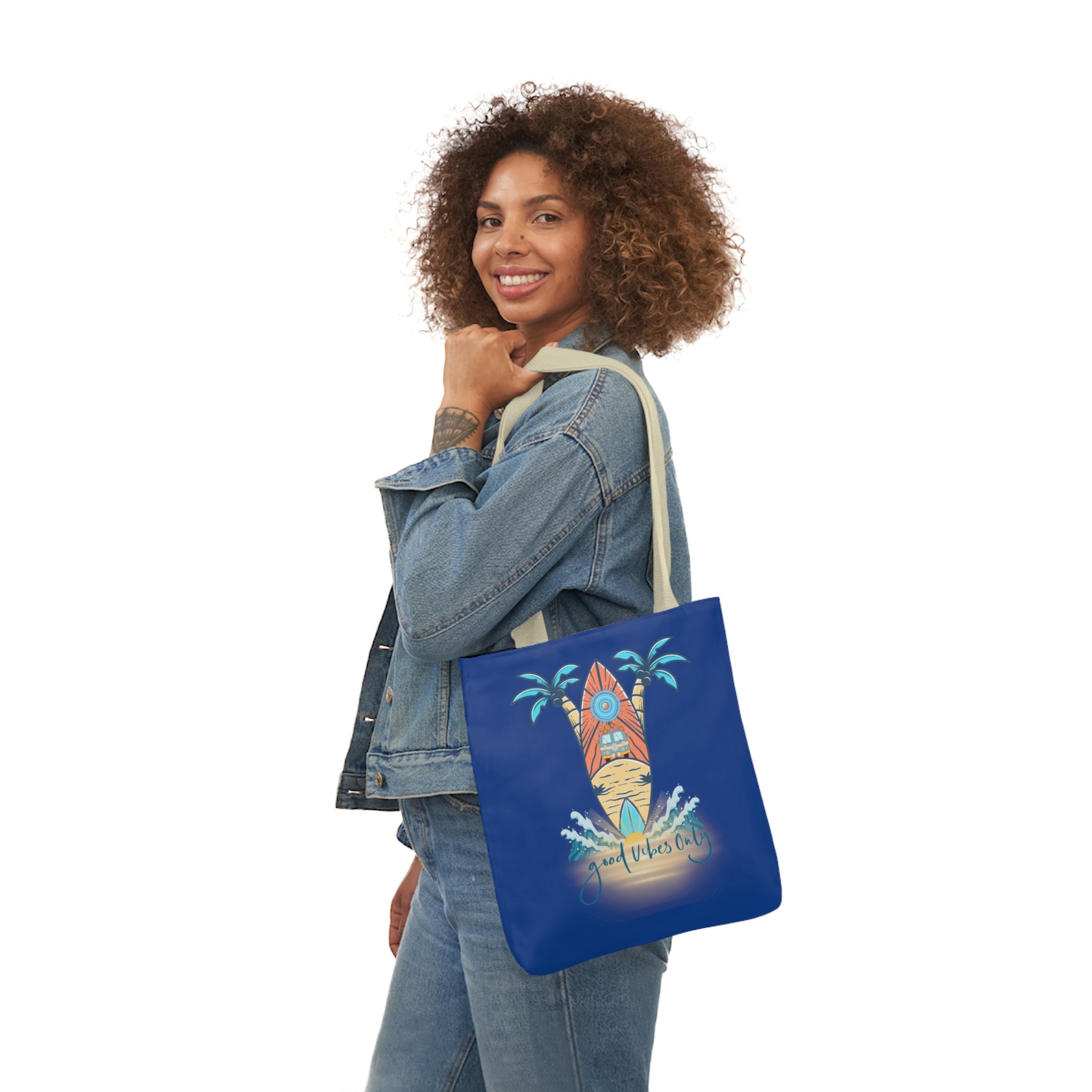 A woman with curly hair wearing a denim jacket and jeans stands sideways, smiling at the camera. She holds a Surf's Up Good Vibes Only Beach Tote by Urban SoulShine & co on her left shoulder, which features surfboards, palm trees, and the text "Good Vibes Only." This polyester canvas bag has comfortable straps perfect for everyday use.