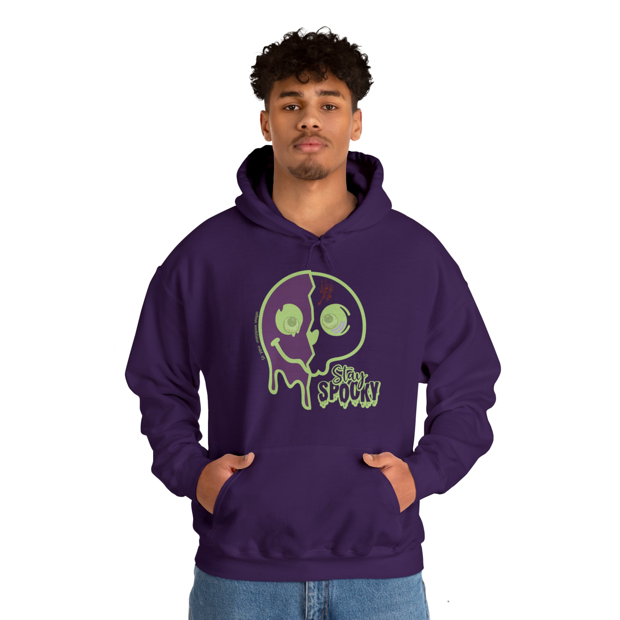 A person stands against a plain white background, wearing a purple hoodie with "Stay Spooky" and a green dripping skull graphic on the front. The person has hands in the hoodie’s front pocket and is looking directly at the camera. They are also wearing jeans from an inspirational wear collection called Soulshine. The hoodie they are wearing is the Stay Spooky Funky Faces Halloween Hoodie (unisex) by Printify.