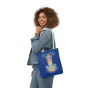 A person with curly hair and a denim jacket is holding a blue tote bag with a colorful surfboard design, palm trees, and waves. The text on the bag reads "Surf's Up Good Vibes Only Beach Tote." The individual, radiating soulshine, is smiling and posing against a plain white backdrop.