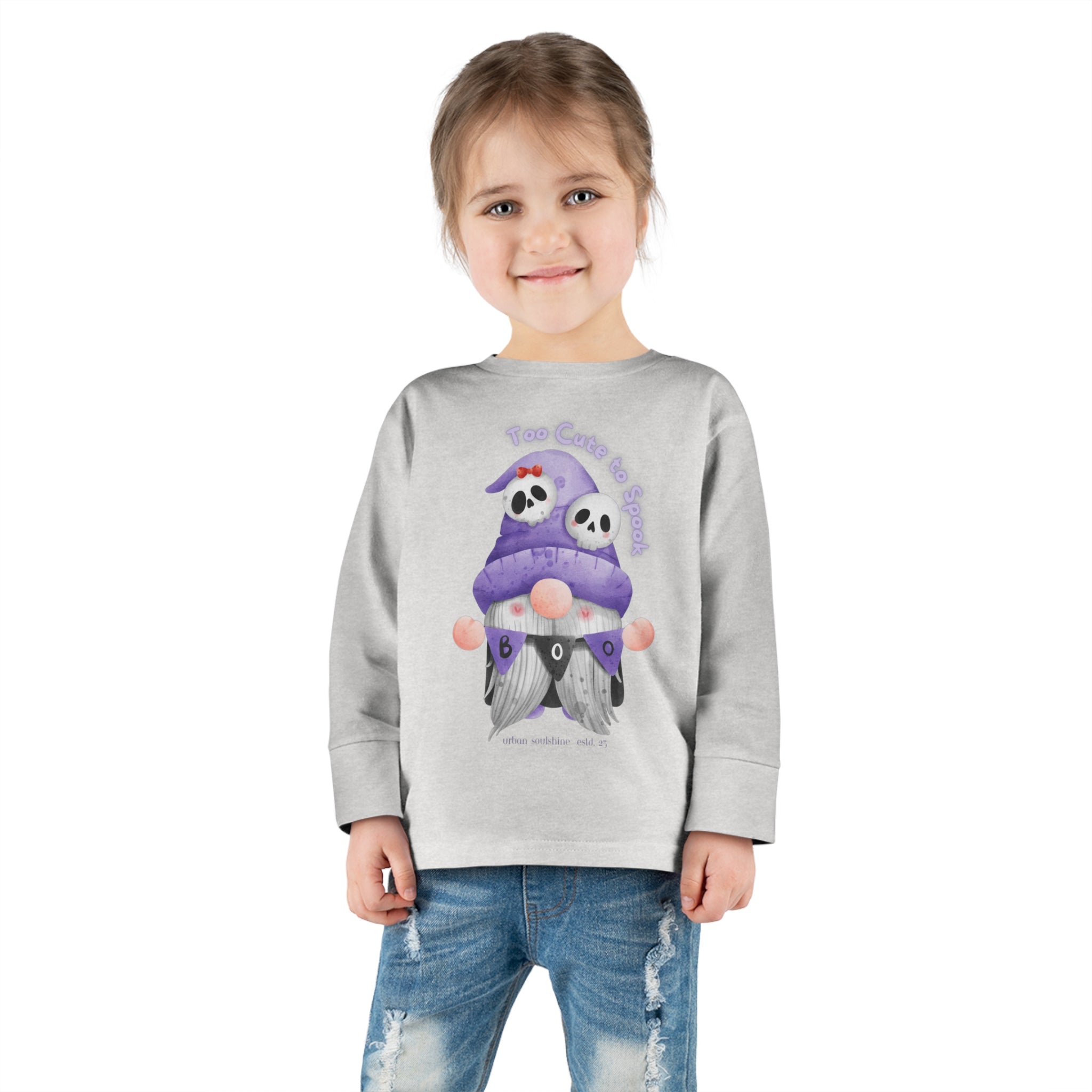 A young child with brown hair stands against a white background, wearing a light grey sweatshirt and blue jeans. The sweatshirt features an illustration of two skeletons seated, and text reading “Too Cute To Spook.” This outfit encourages you to express yourself with a touch of whimsy. The long sleeve tee is the Too Cute To Spook Halloween Toddler Long Sleeve Tee (unisex Tot) by Printify.