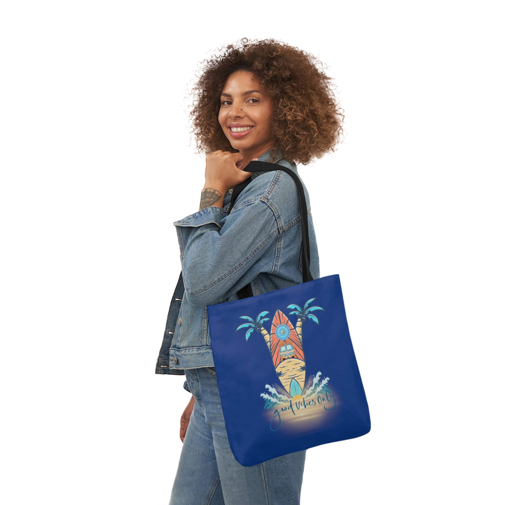 A person with curly hair wearing a denim jacket holds a blue Surf's Up Good Vibes Only Beach Tote by Printify over their shoulder, showcasing a colorful surfboard design with palm trees and text that reads "Just ride the wave." The scene radiates soulshine and artistic expression.