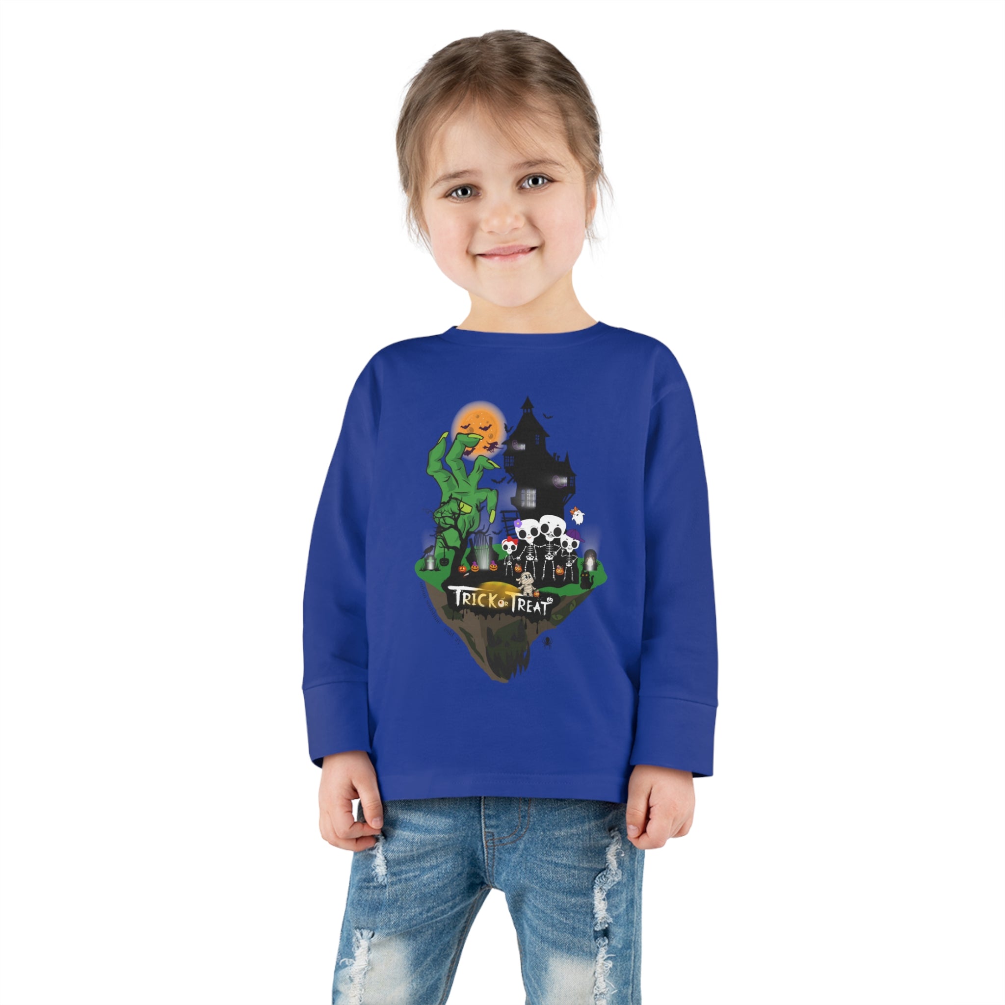 A young girl with a ponytail is wearing the Spooky Island Halloween Toddler Long Sleeve Tee by Urban SoulShine & Co. This 100% combed ringspun cotton shirt features a Halloween-themed design with the text "Trick Or Treat" and illustrated characters. She is smiling and standing with her hands by her sides, also sporting distressed jeans.